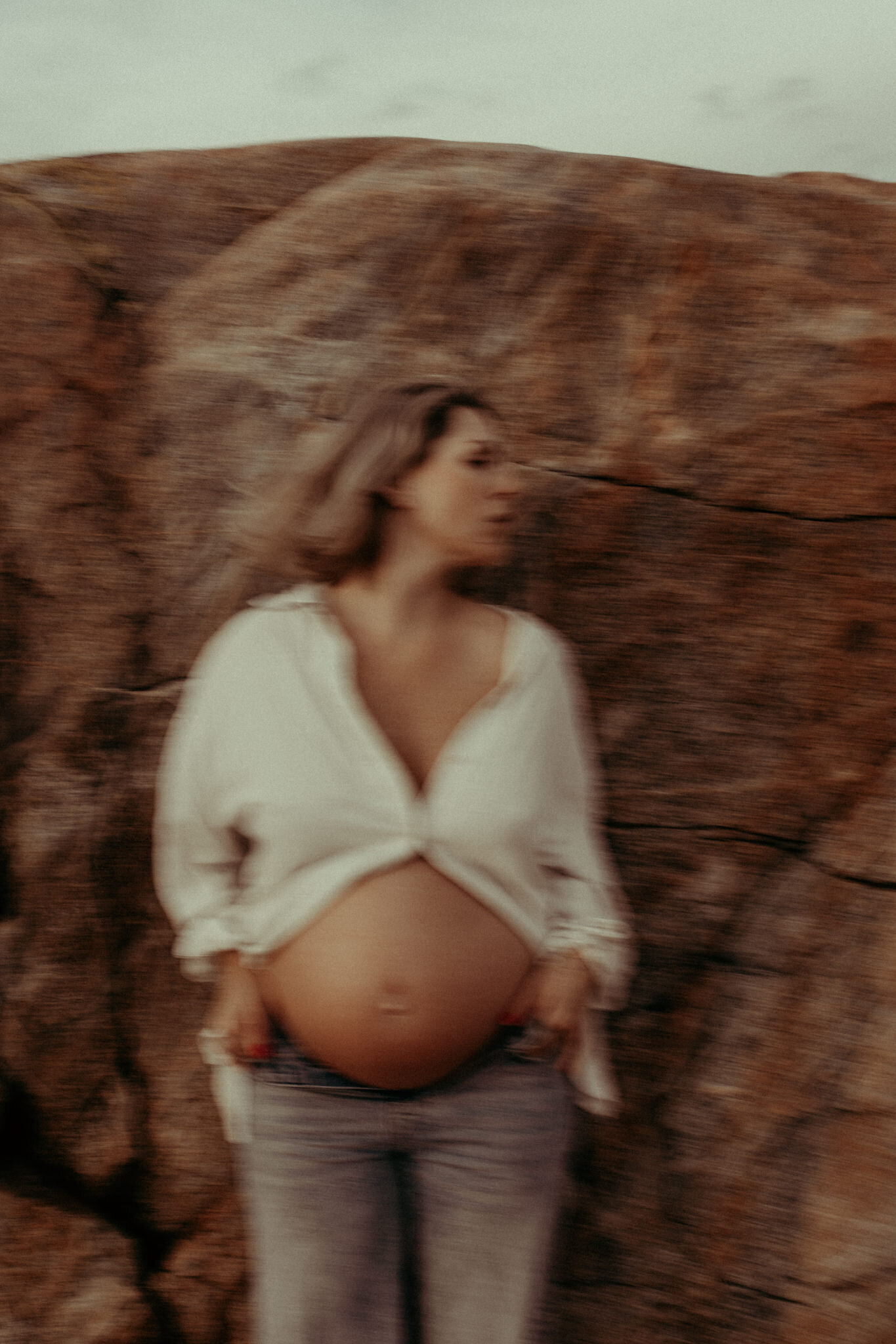 en gravid mamma i vit skjorta och jeans lutar sig mot klippor