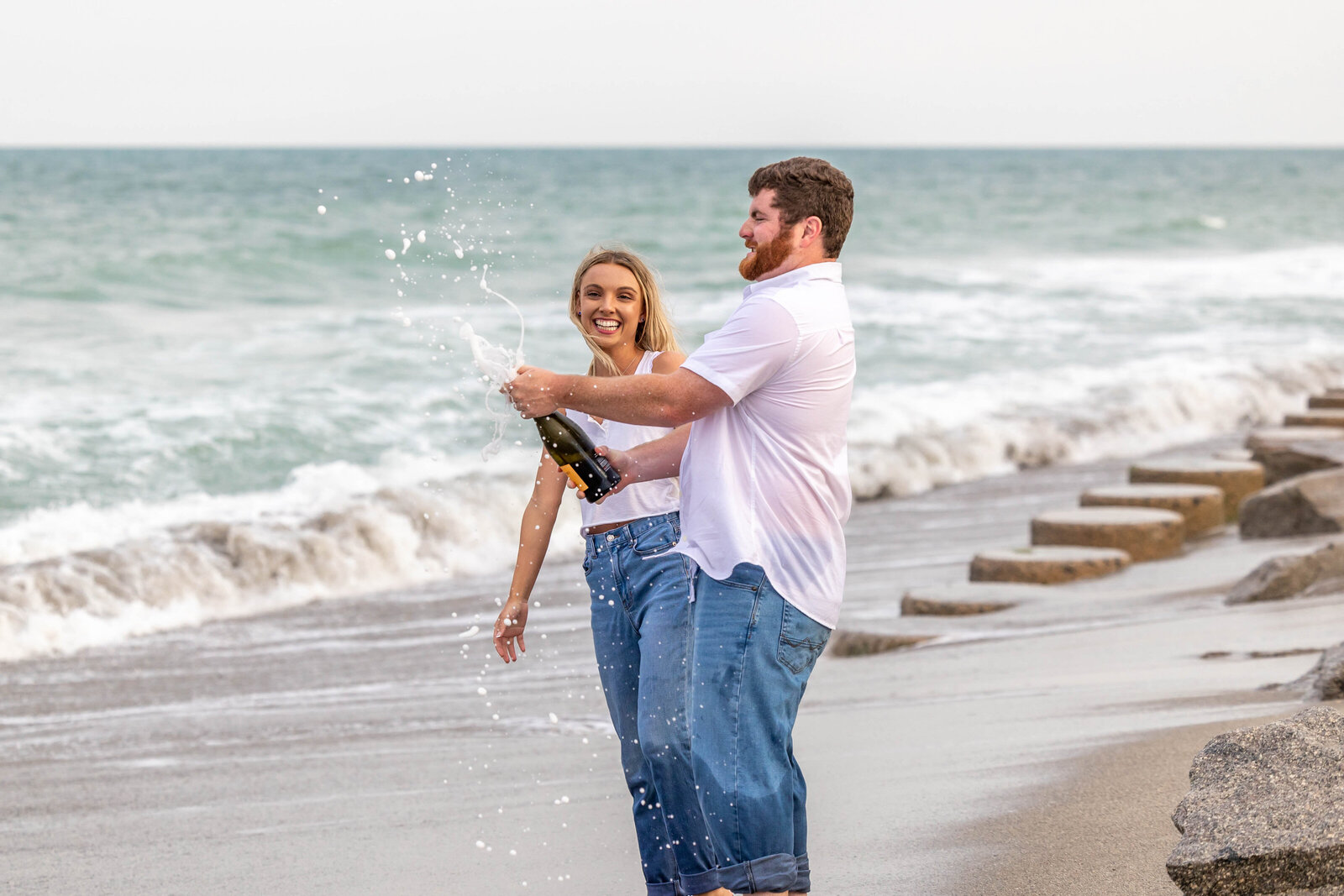 A couple joyfully popping champagne on the beach in Fort Fisher, NC, with waves in the background. This candid moment reflects the excitement of engagement, showcasing creative wedding videography and photography for couples in North Carolina.