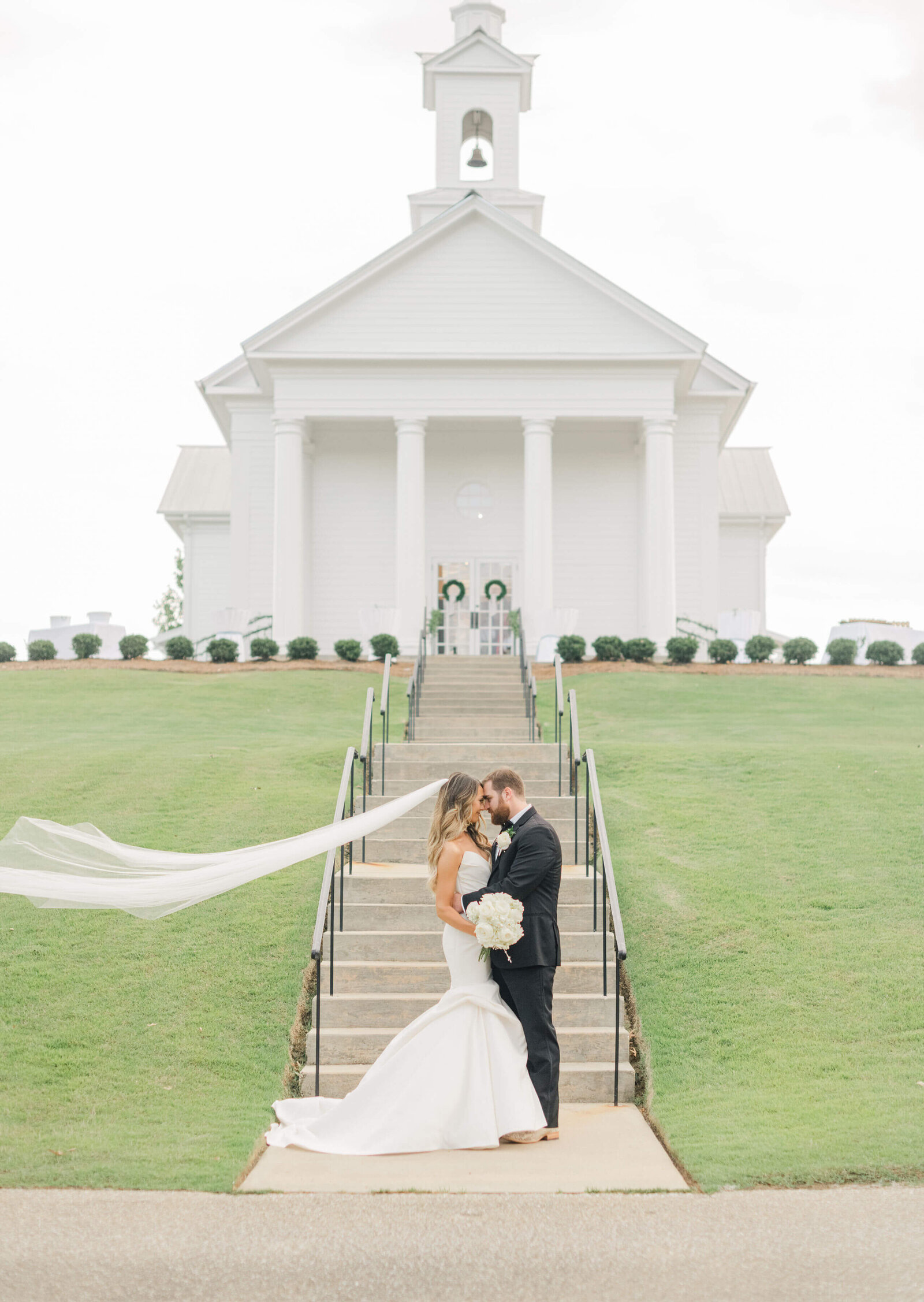 Bride and groom posing closely with bride veil flying in front of a white church in Alabama