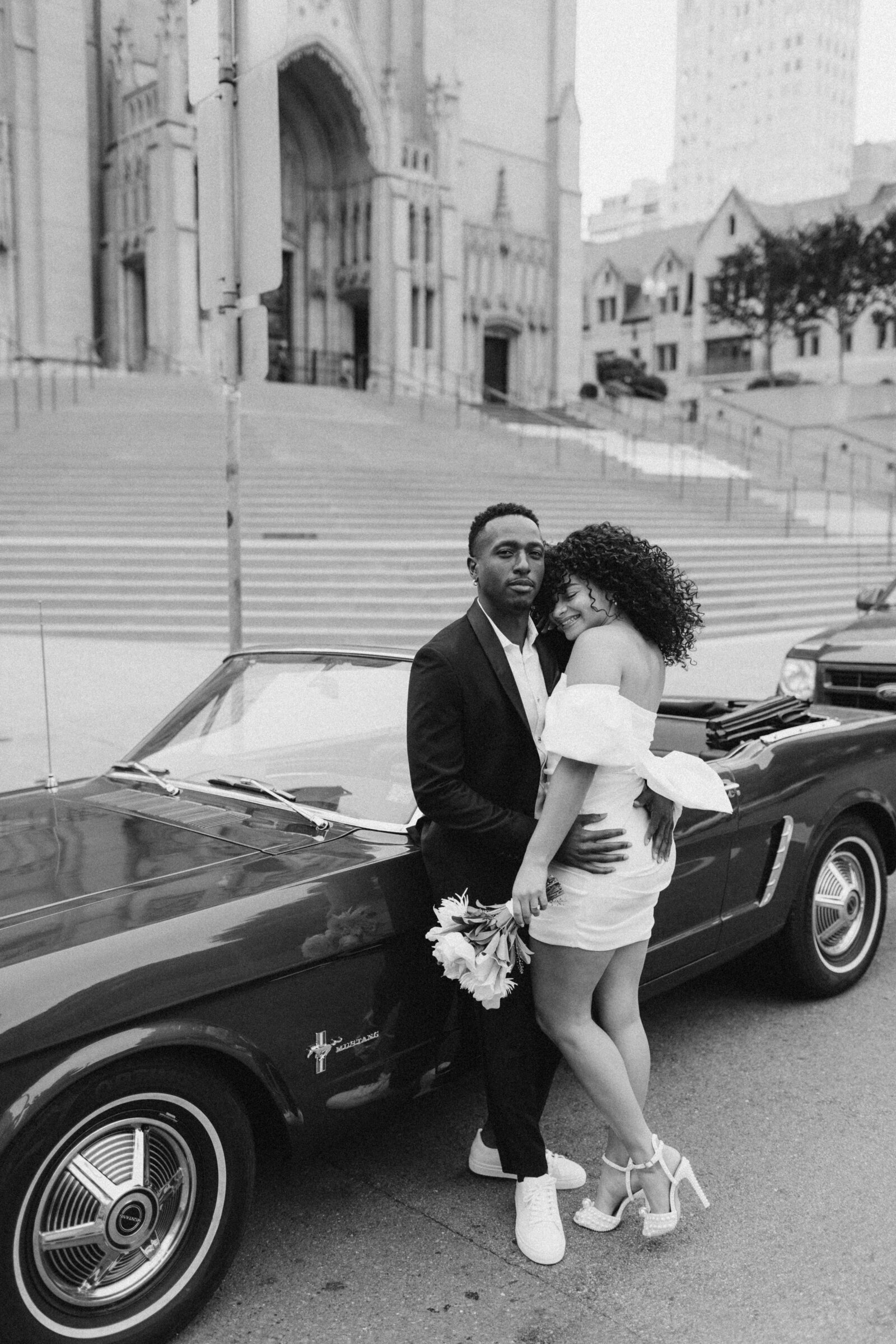 A couple dressed in formal wear poses for a engagement photoshoot in San Francisco beside a classic car, with a cathedral staircase in the background. The woman holds a bouquet, leaning into the man affectionately.