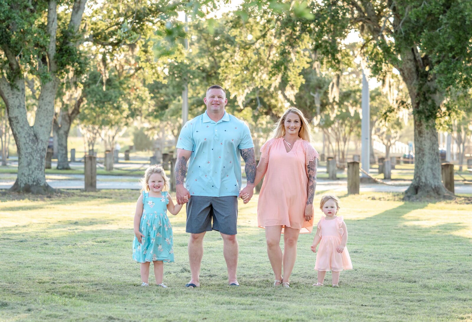 A family of four stands hand in hand in a sunlit park near Wilmington, NC. The parents and their two young daughters are dressed in pastel summer outfits, surrounded by lush green trees. Ideal for those seeking artistic family photography in Wilmington, NC.