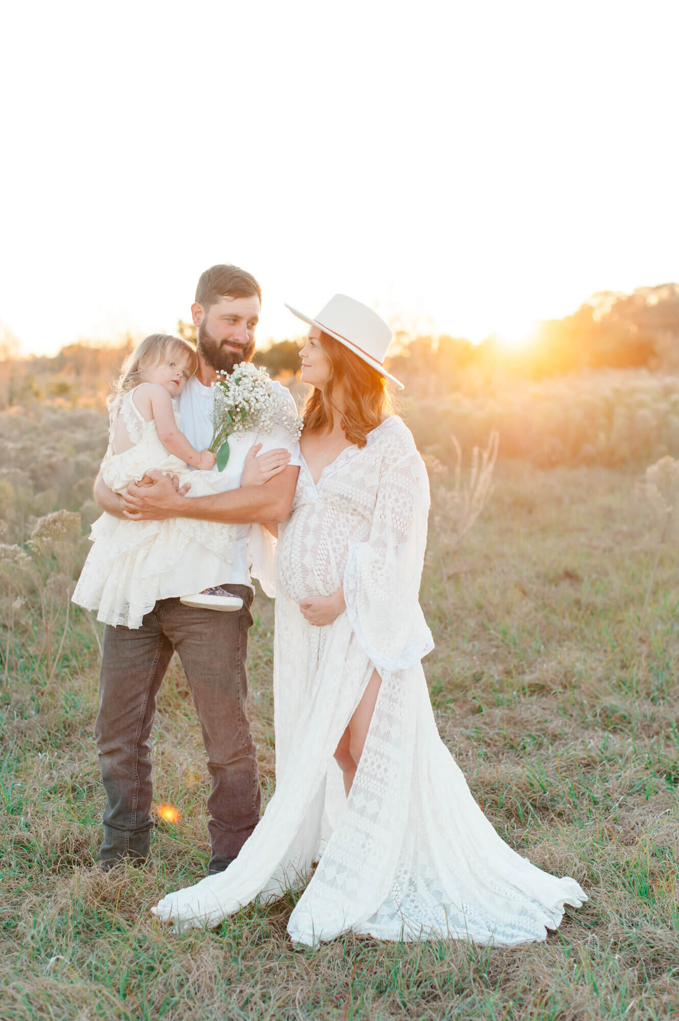 Pregnant couple standing in a tall grass field holding their young daughter at sunset
