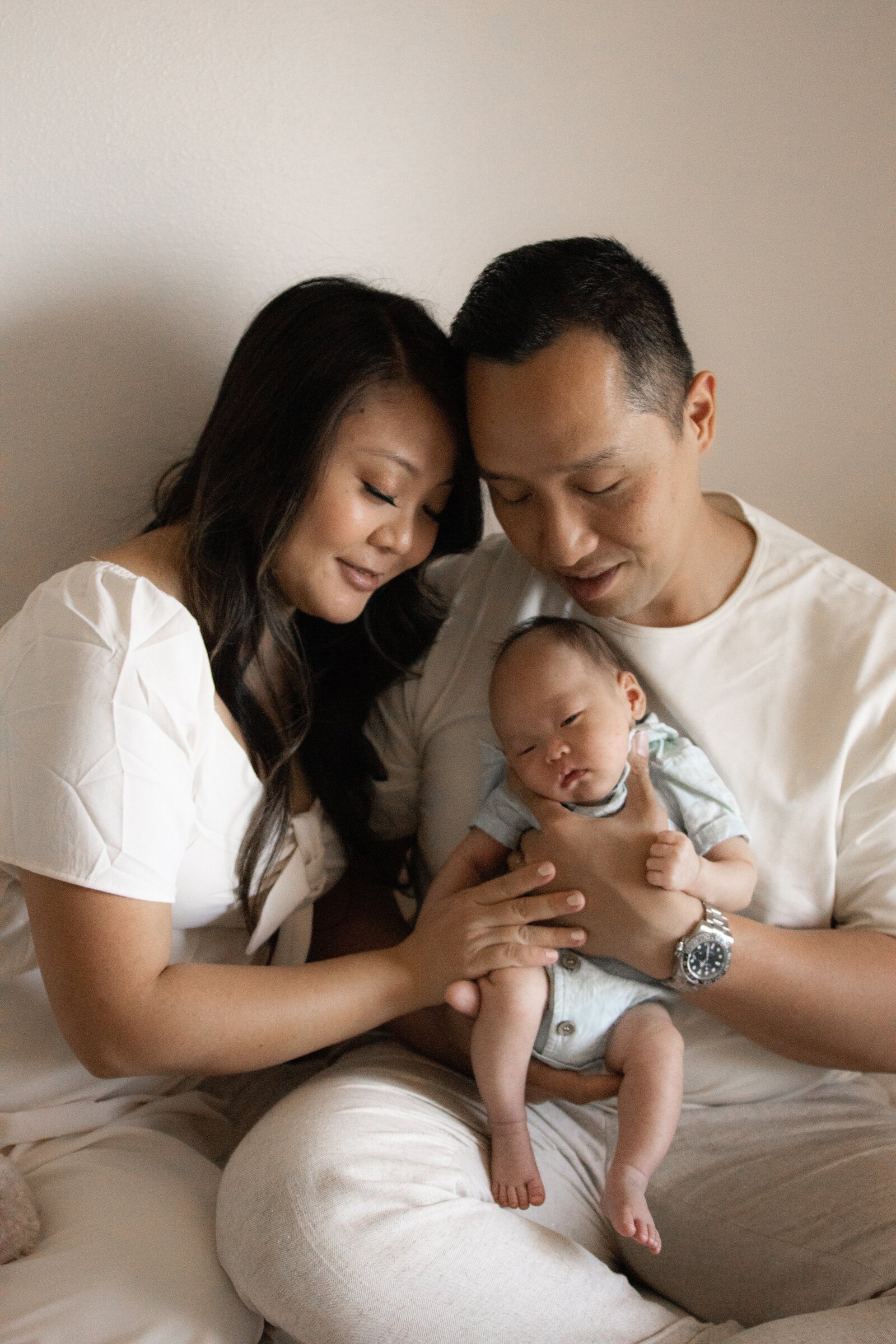 Dad braces sleeping newborn son against chest while cuddled up with mom on bed.