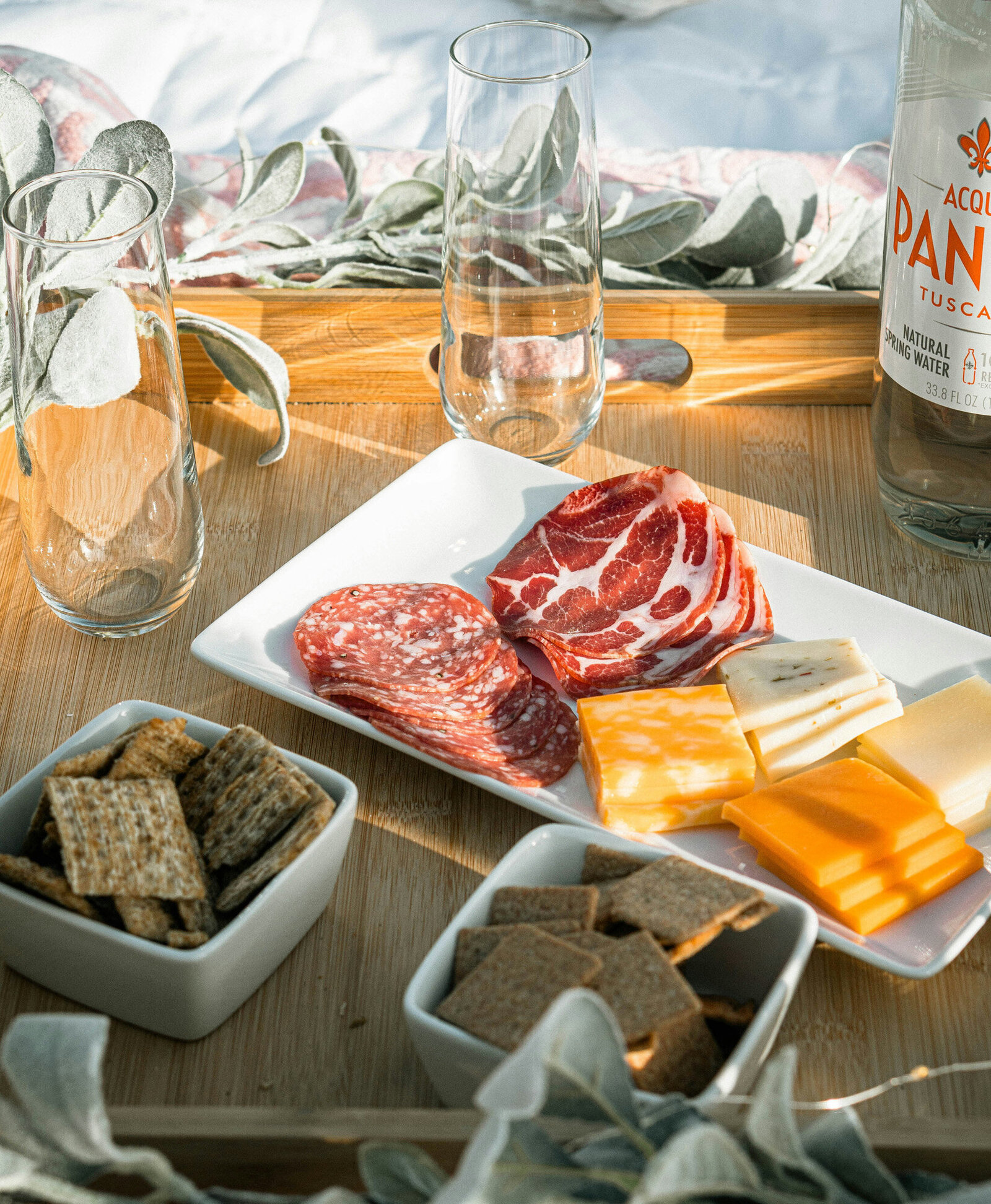 wooden tray holds two empty glasses, a bottle of sparkling water, and a white rectangular plate with assorted meats and cheeses and two small bowls nearby that has crackers