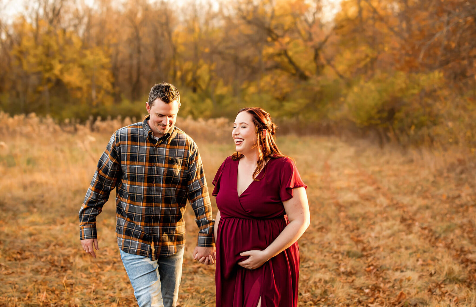 A smiling couple walks hand in hand along a grassy path in autumn. Captured by a Bloomington maternity photographer, the mom-to-be cradles her baby bump as fall foliage surrounds them—perfect for heartfelt pregnancy photography and timeless maternity portraits.