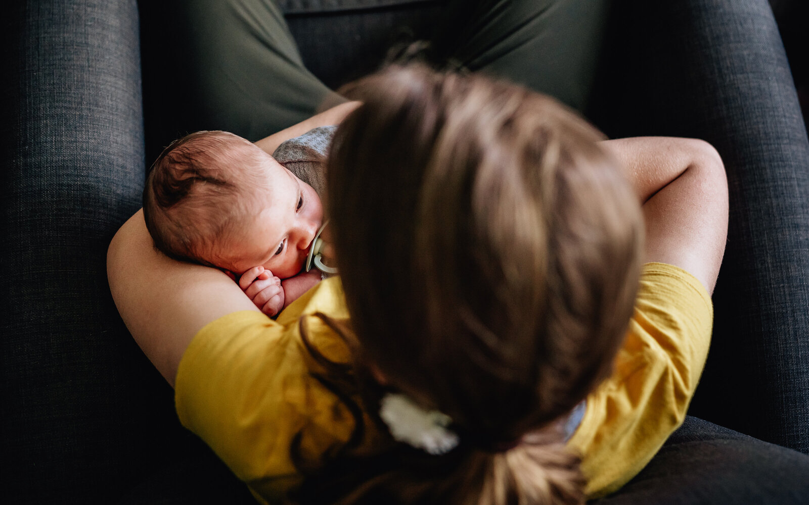 A mother cradles her newborn baby in her arms while sitting in a chair. The baby is wrapped in a blanket and gently sucking on a pacifier.