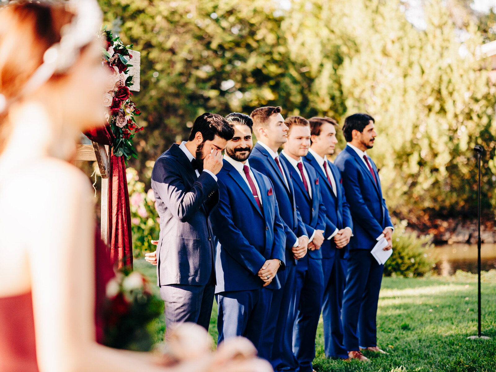 A group of groomsmen in blue suits stand outdoors at a wedding ceremony, one man covering his face emotionally. Captured by a San Antonio Wedding Photographer, trees and greenery fill the background while a bridesmaid is blurred in the foreground.



