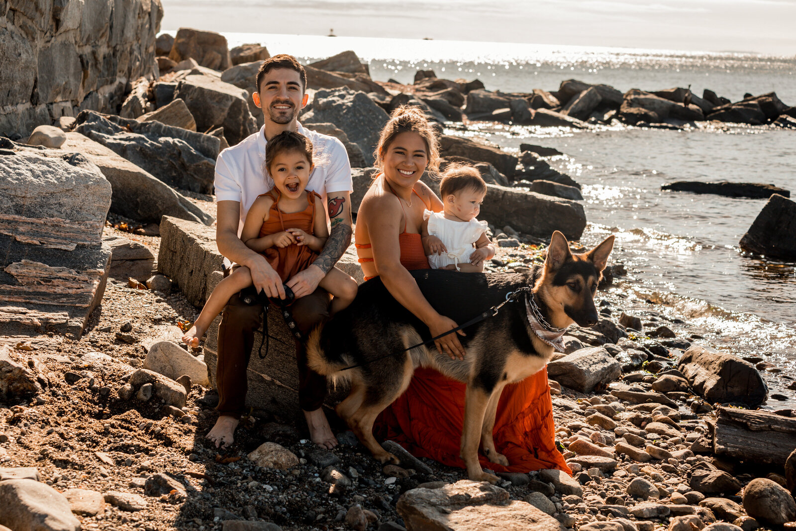 family smiling and sitting on rocks with their dog on the beach