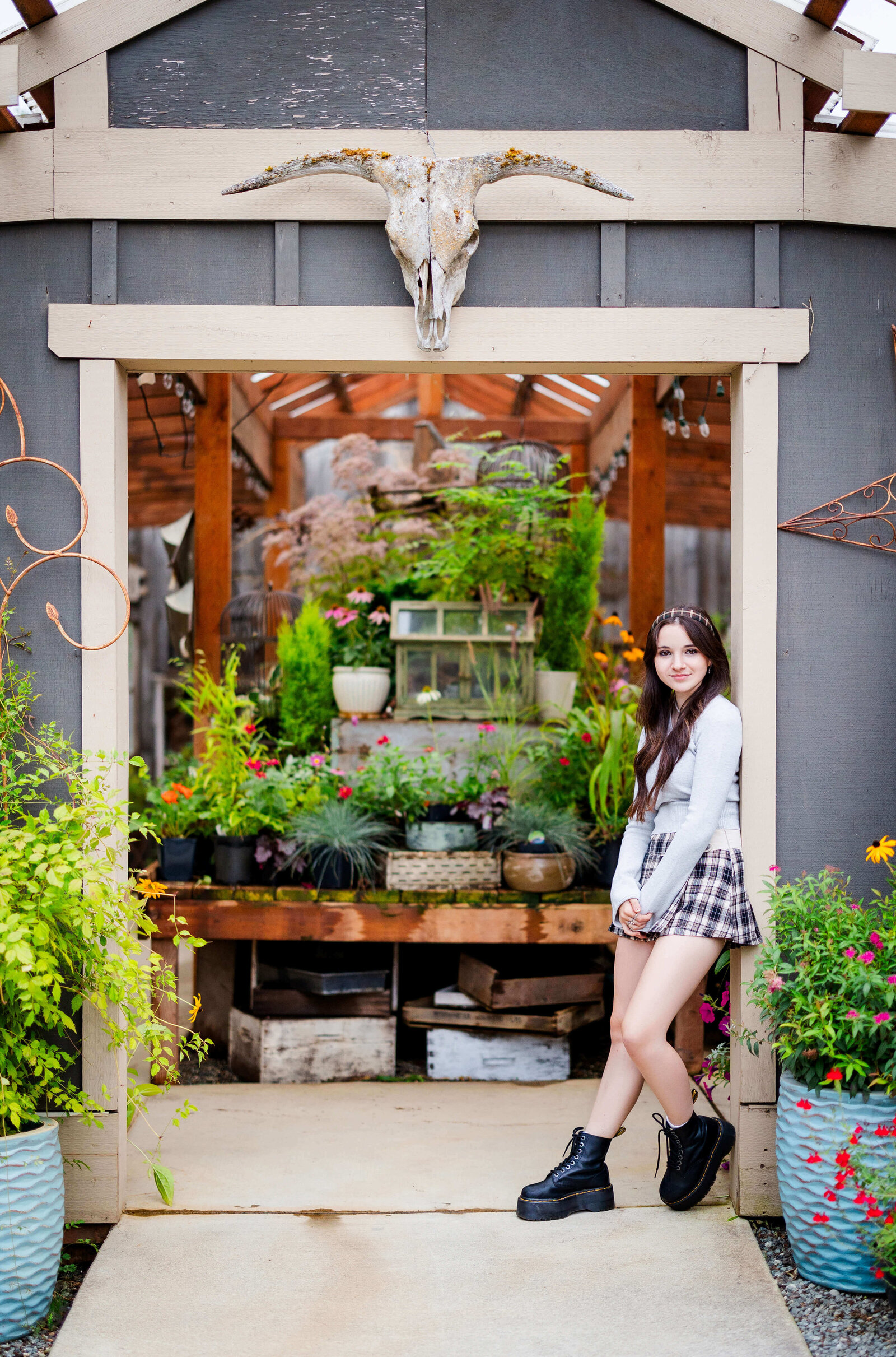 A teenage girl in a plaid skirt leans on the entrance to a greenhouse