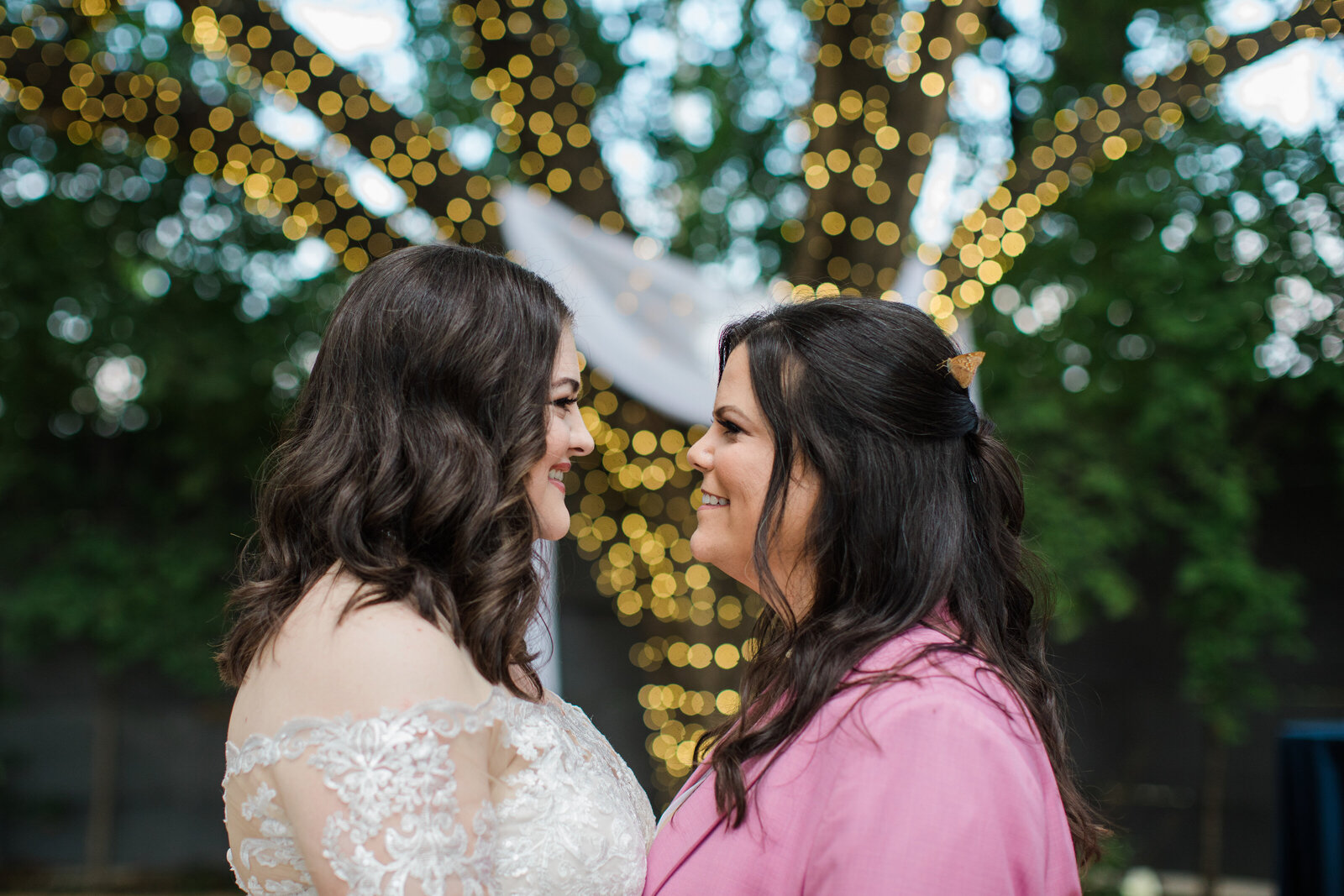 Two brides look at each other in front of a big oak tree covered in twinkle lights at Artspace111 venue in Fort Worth, Texas. The bride on the left is wearing a long sleeve lace white dress. The bride on the right is in a pink suit. They both have brown hair, and the bride with the pink suit has an orange monarch butterfly in her hair.