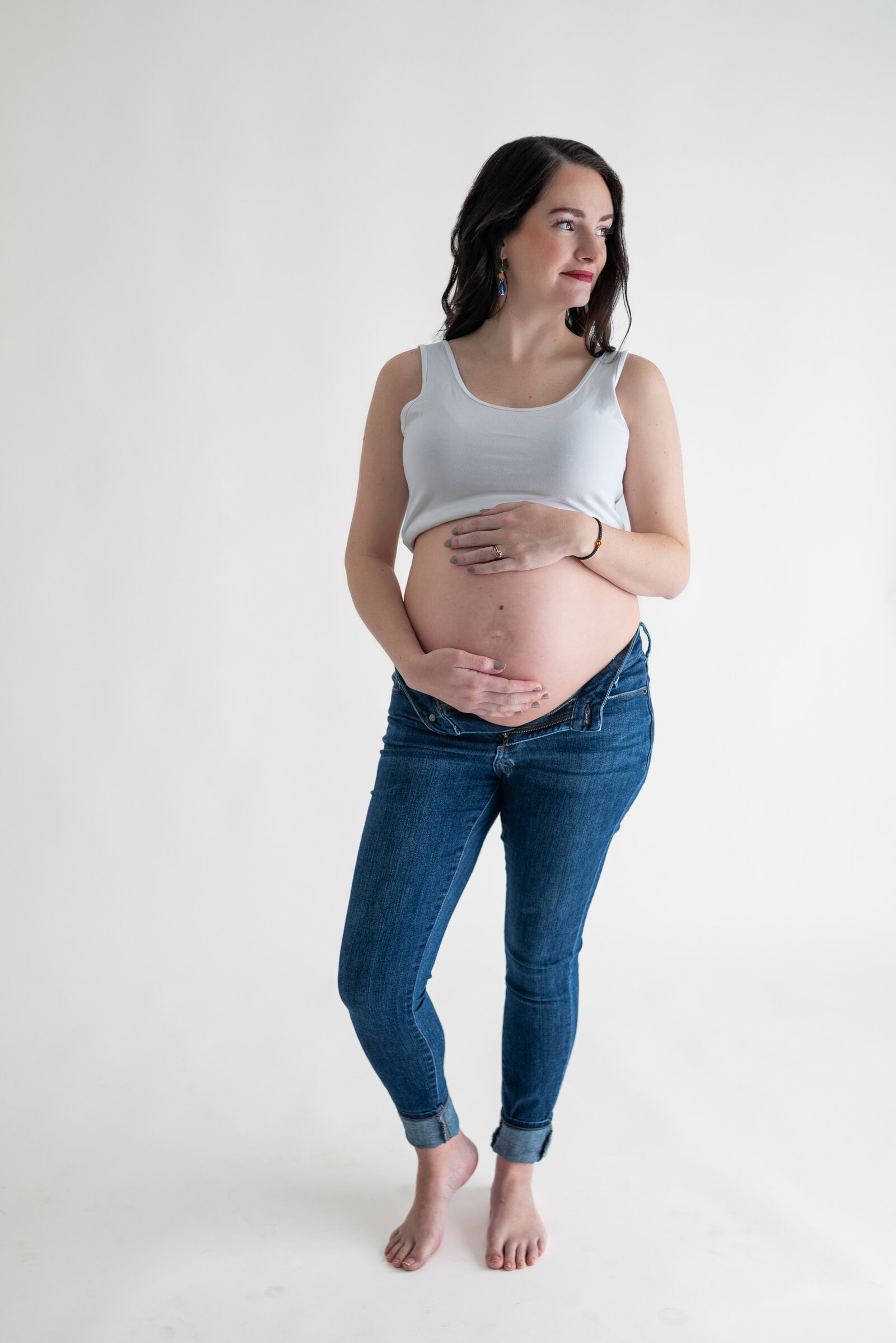 A pregnant woman in a tank top and jeans and holding her stomach while posing for her maternity photos in Huntsville Alabama