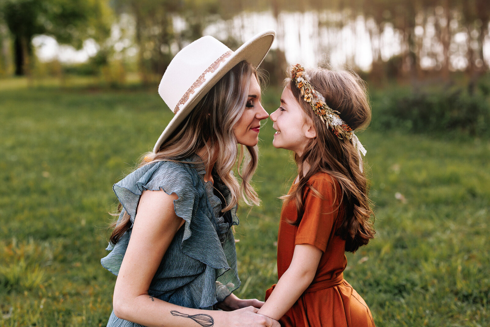 A mother dressed in a dusty blue ruffled dress and a cream large brim hat, nuzzles her daughter dressed in a rust dress and floral crowns nose