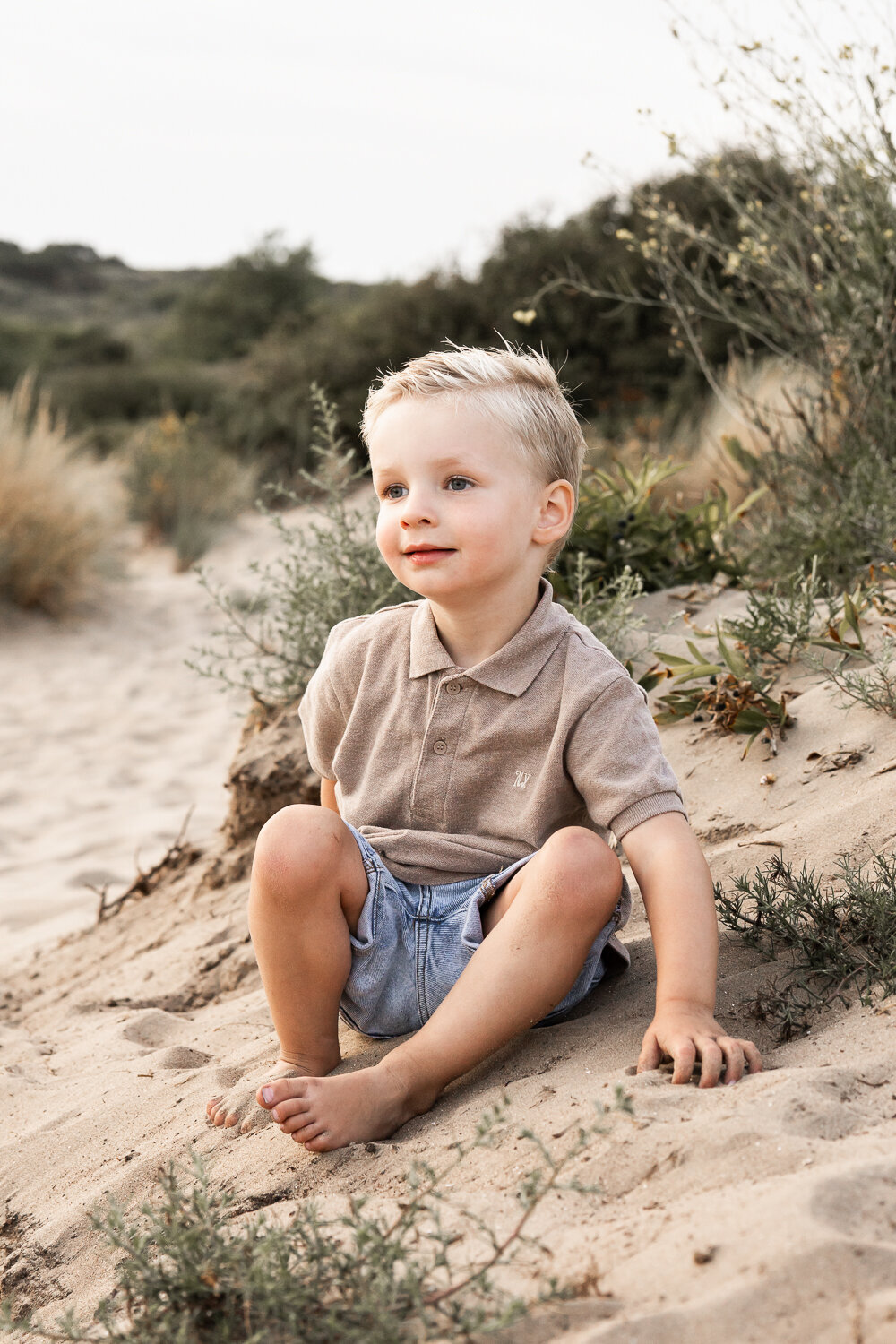 Een peuter zit met een ontspannen glimlach in de duinen tijdens een fotoshoot met zijn gezin.
