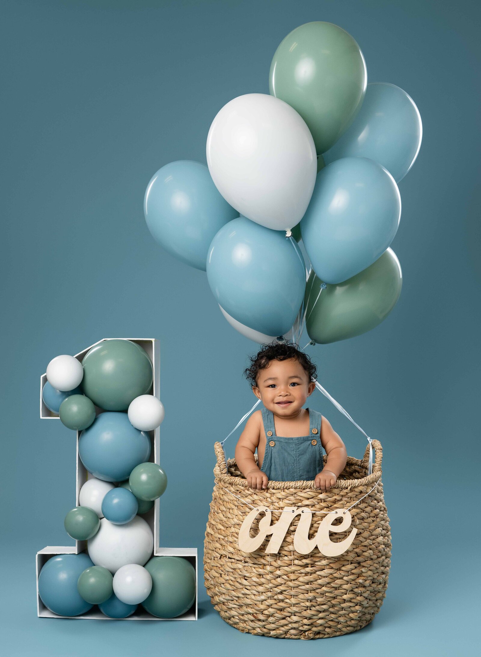 1 year old taking birthday photos, standing in balloon basket smiling.