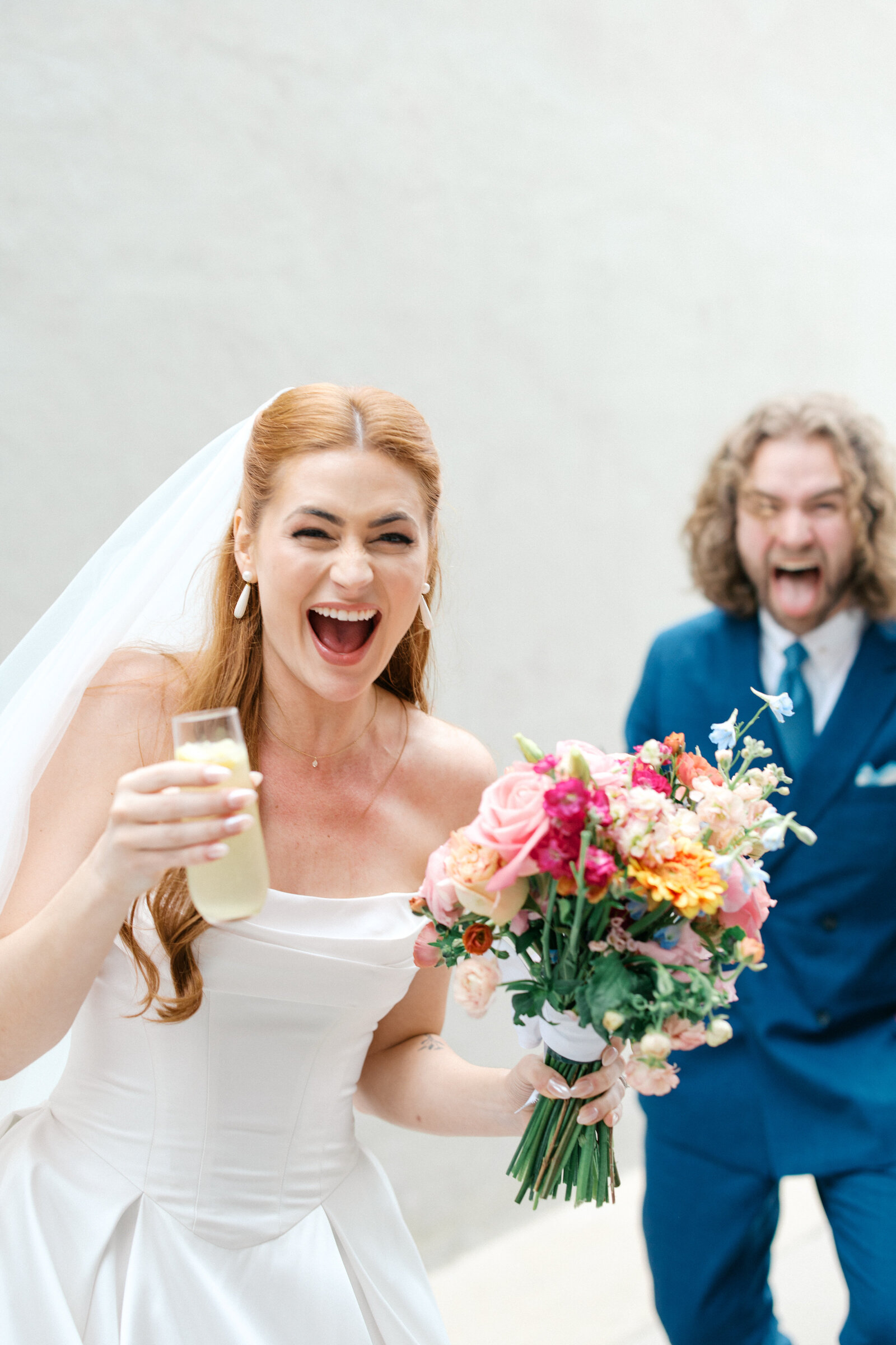 Bride laughs with yellow cocktail and colorful bouquet with groom behind her laughing and sticking tongue out 