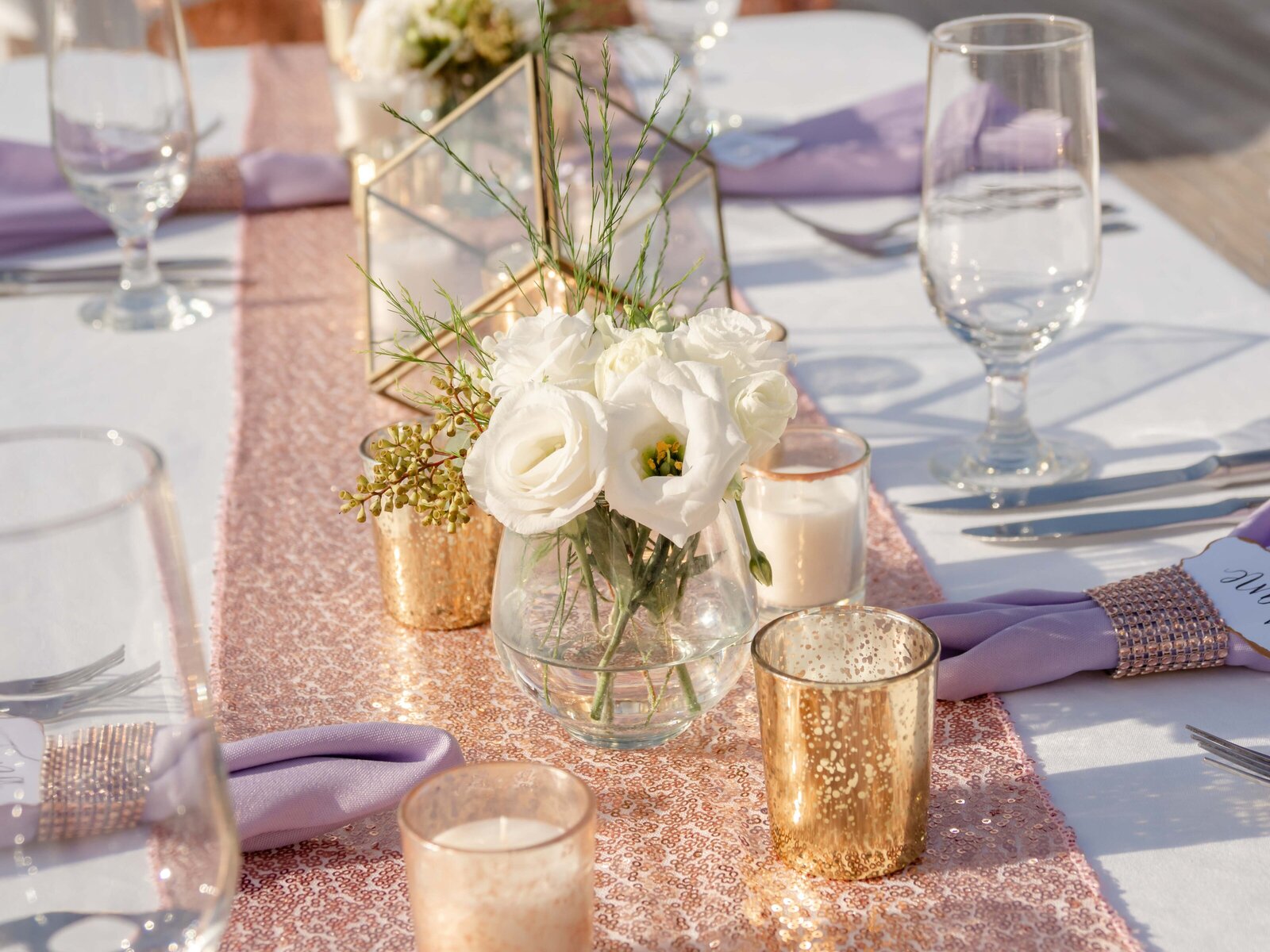 A beautifully decorated wedding table featuring a rose gold sequin runner, white florals, lavender napkins, and gold candle votives, captured during a Wilmington, NC wedding, blending modern elegance and romantic details.