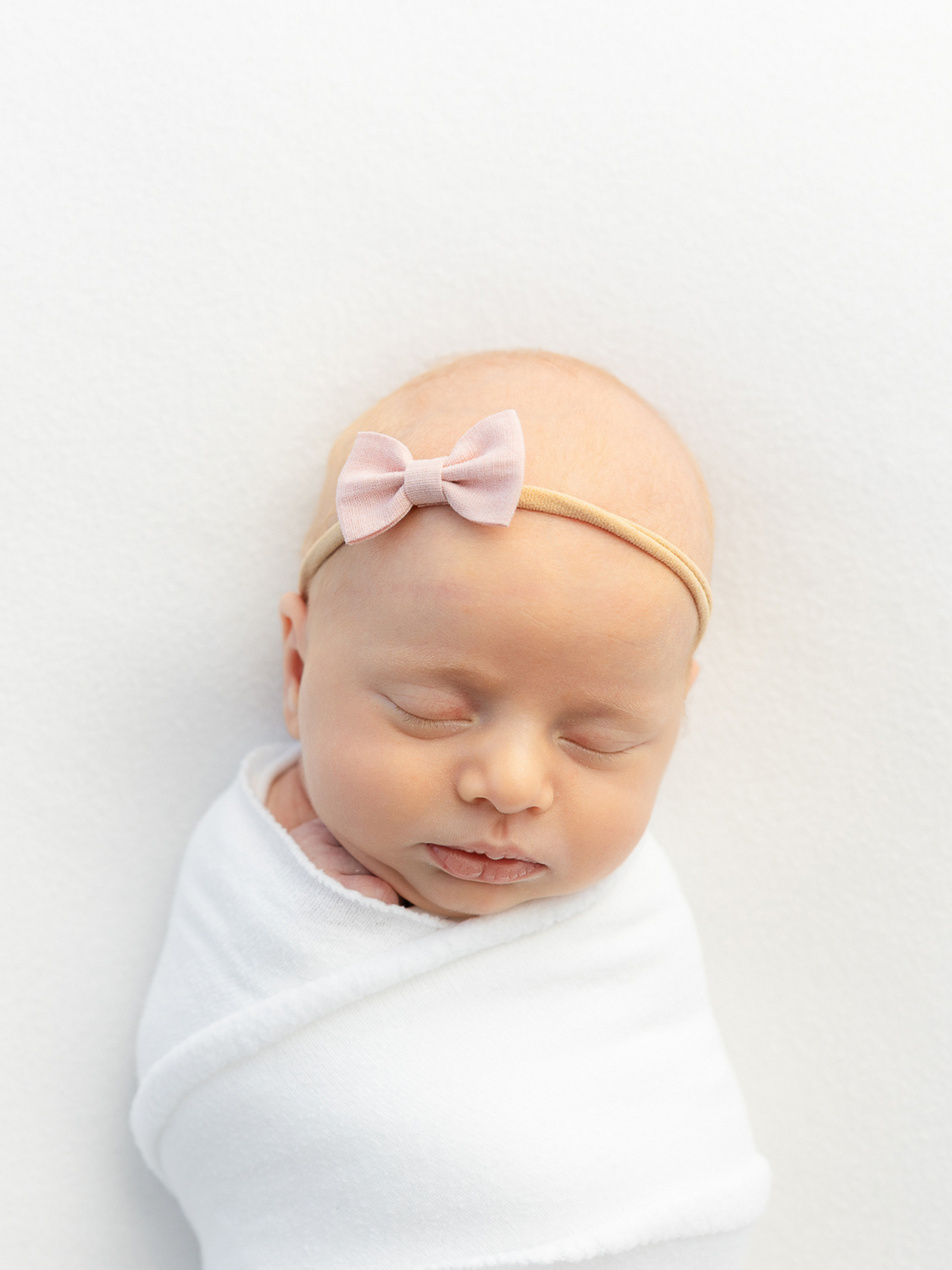 sleeping newborn baby girl wearing simple pink bow is wrapped gently in a white swaddle