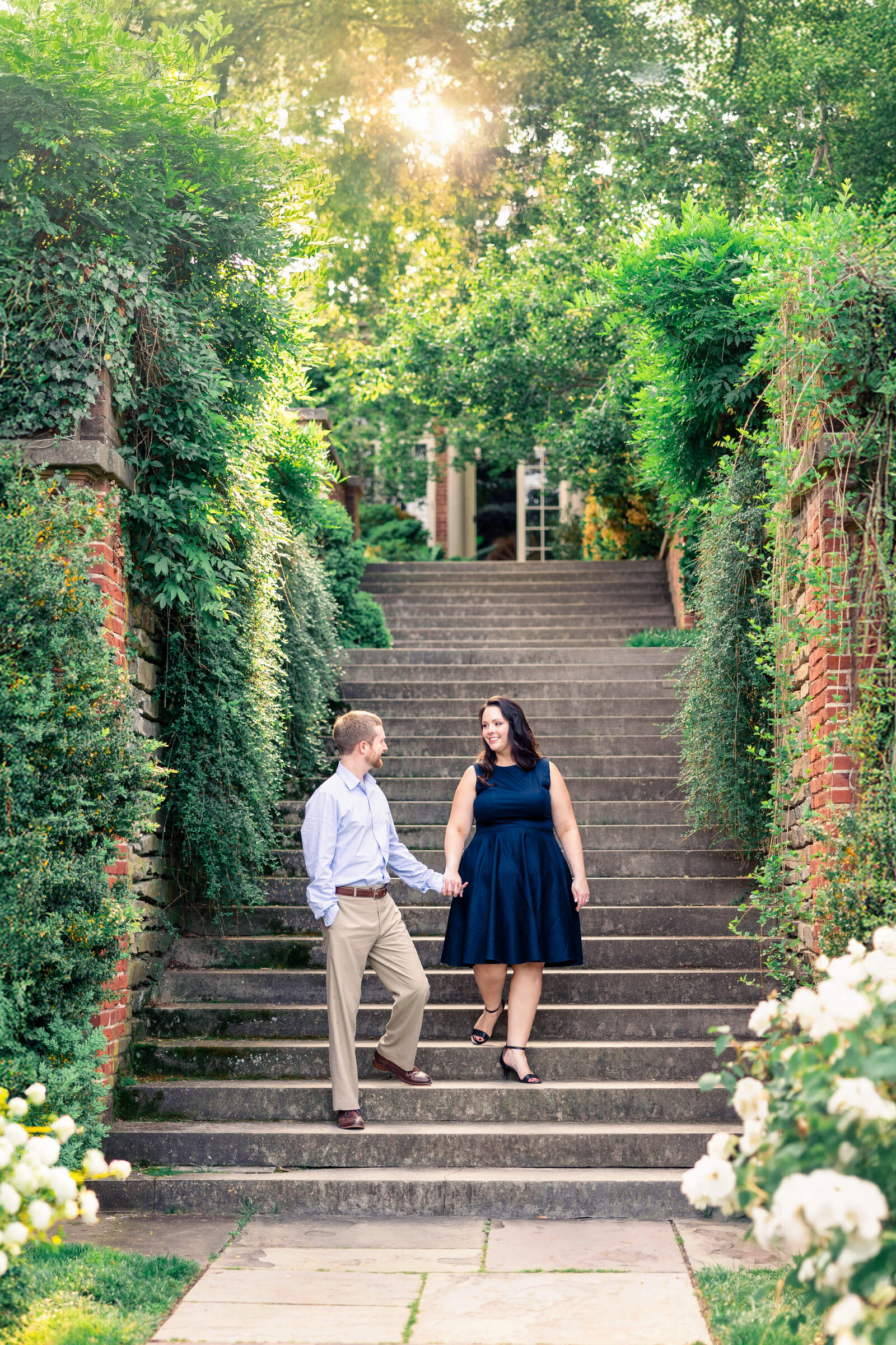 A couple is walking down stone steps surrounded by lush greenery and blooming flowers. The sun filters through the trees above, casting a warm glow over the scene. Both are smiling and holding hands, creating a joyful and romantic atmosphere.