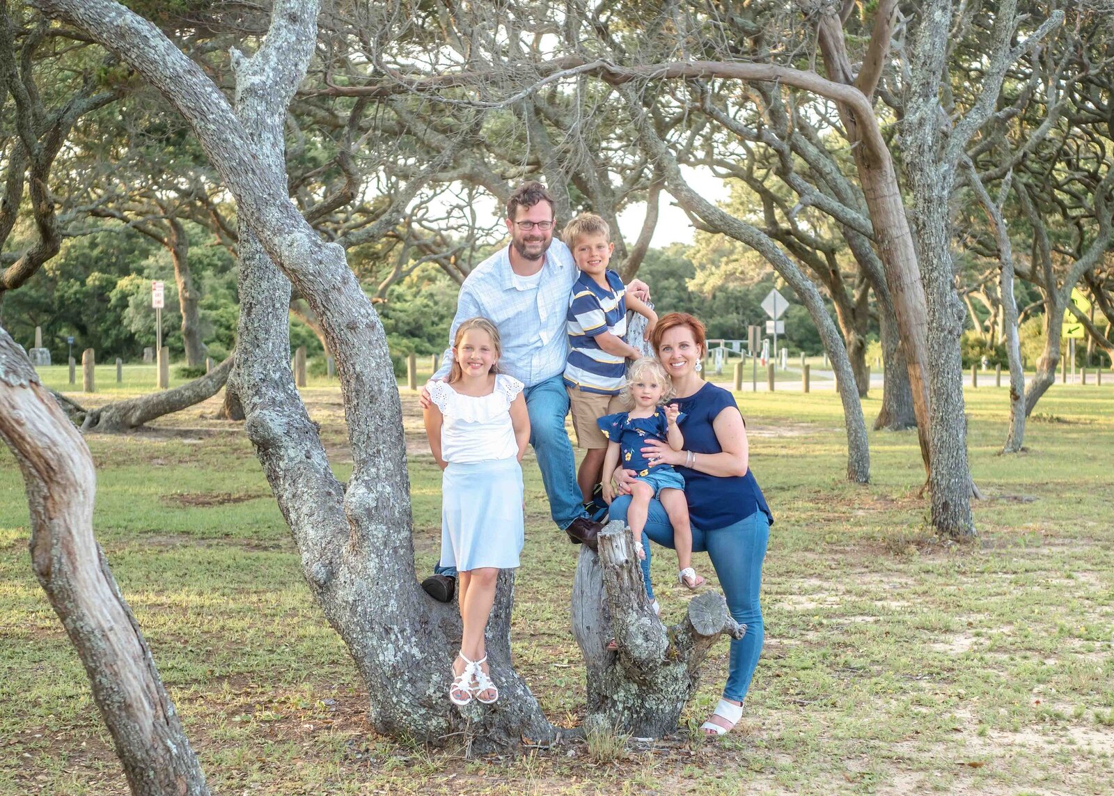 A family of five poses among the trees in a Wilmington park. The parents and their three children are dressed in coordinated blue and white outfits, enjoying the natural setting. This image is ideal for destination family photographer NC, capturing candid family joy.