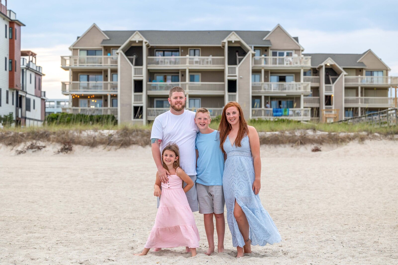 A family of four poses on the sandy beach with beachfront condos in the background. The parents and children are dressed in pastel outfits, capturing a relaxed and joyful vacation vibe. Ideal for North Carolina family photography, highlighting beachside living.