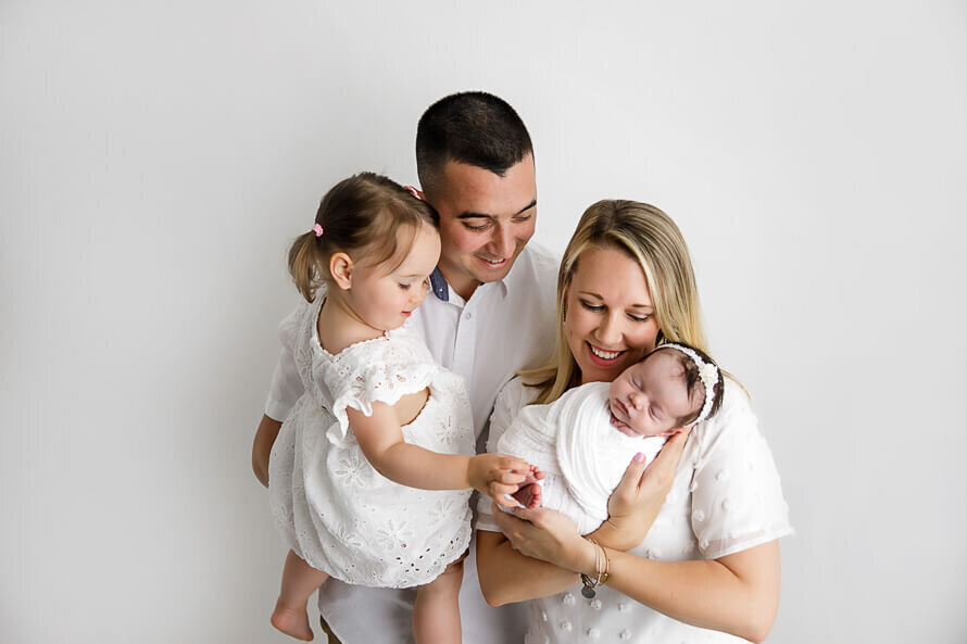 New family of four look at their brand new baby in a white photography studio.