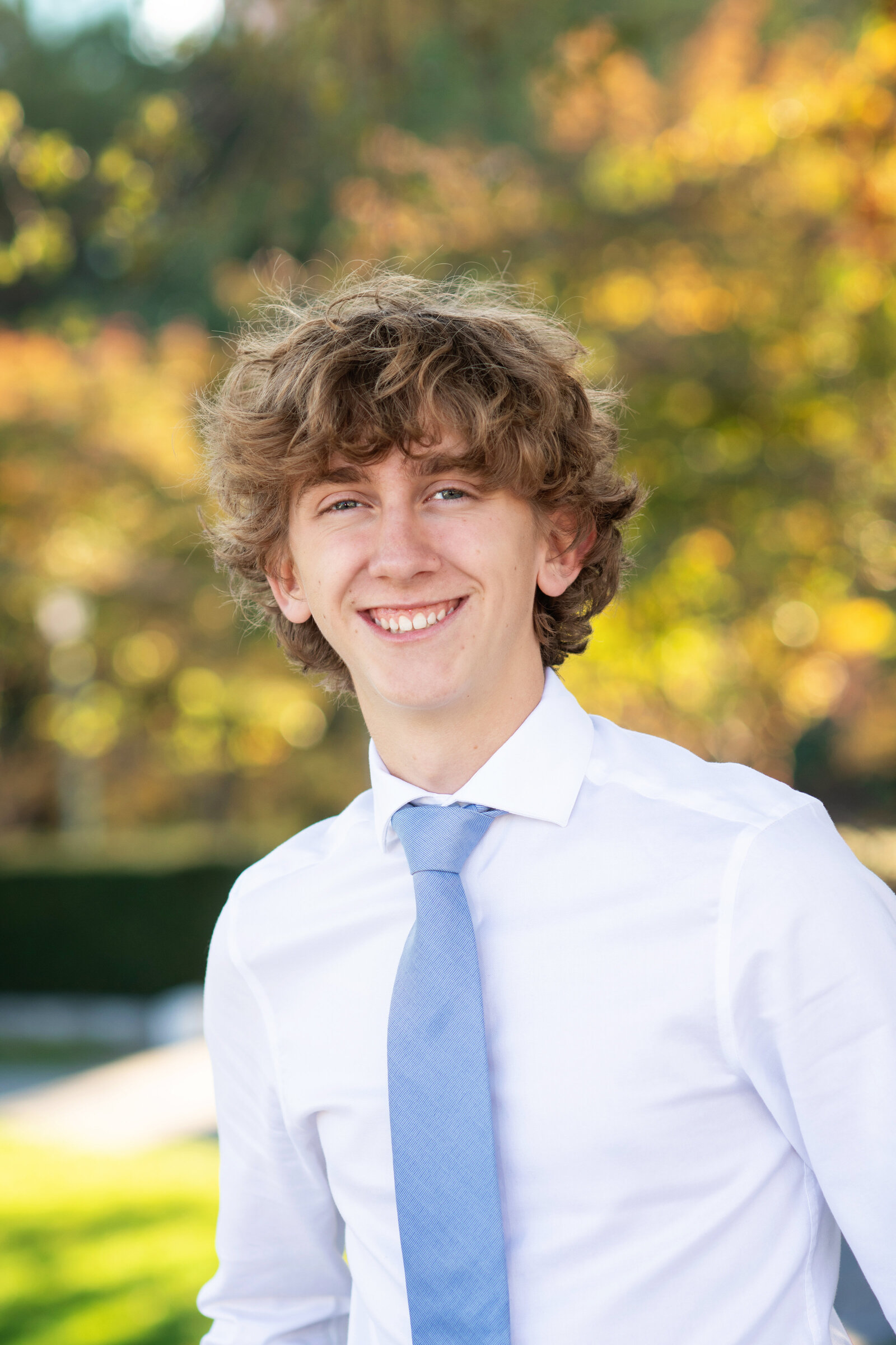 smiling teenager poses in shirt and tie with fall leaves in the background