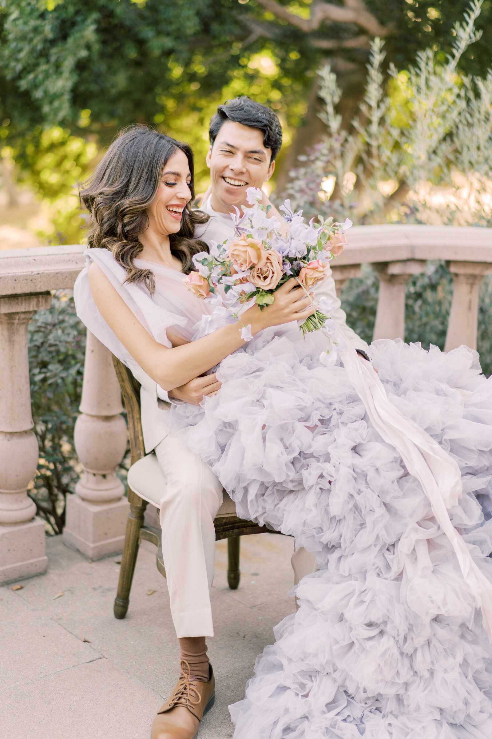 Portrait of bride and groom in a lavender gown and cream suit sitting on a chair outdoors with a bouquet.