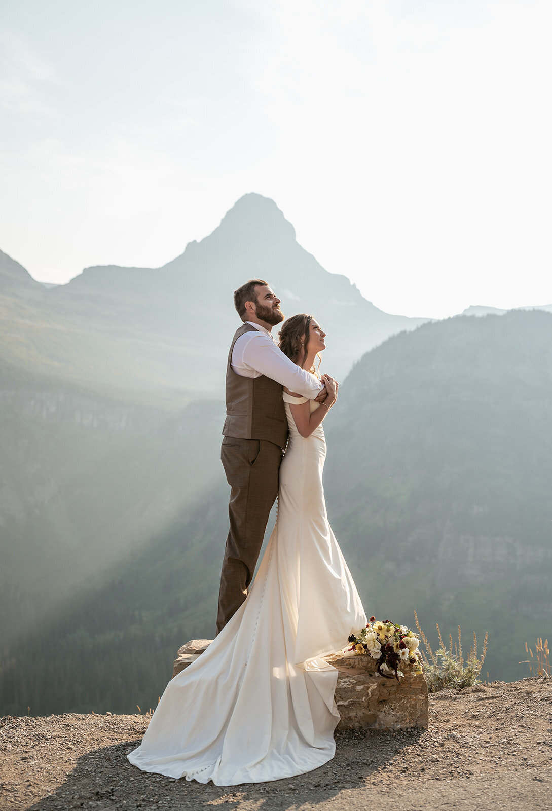 A groom embraces his bride from behind as they stand on a sunlit cliff with mountain peaks glowing in the distance during their Glacier National Park elopement, captured by Sydney Breann Photography.