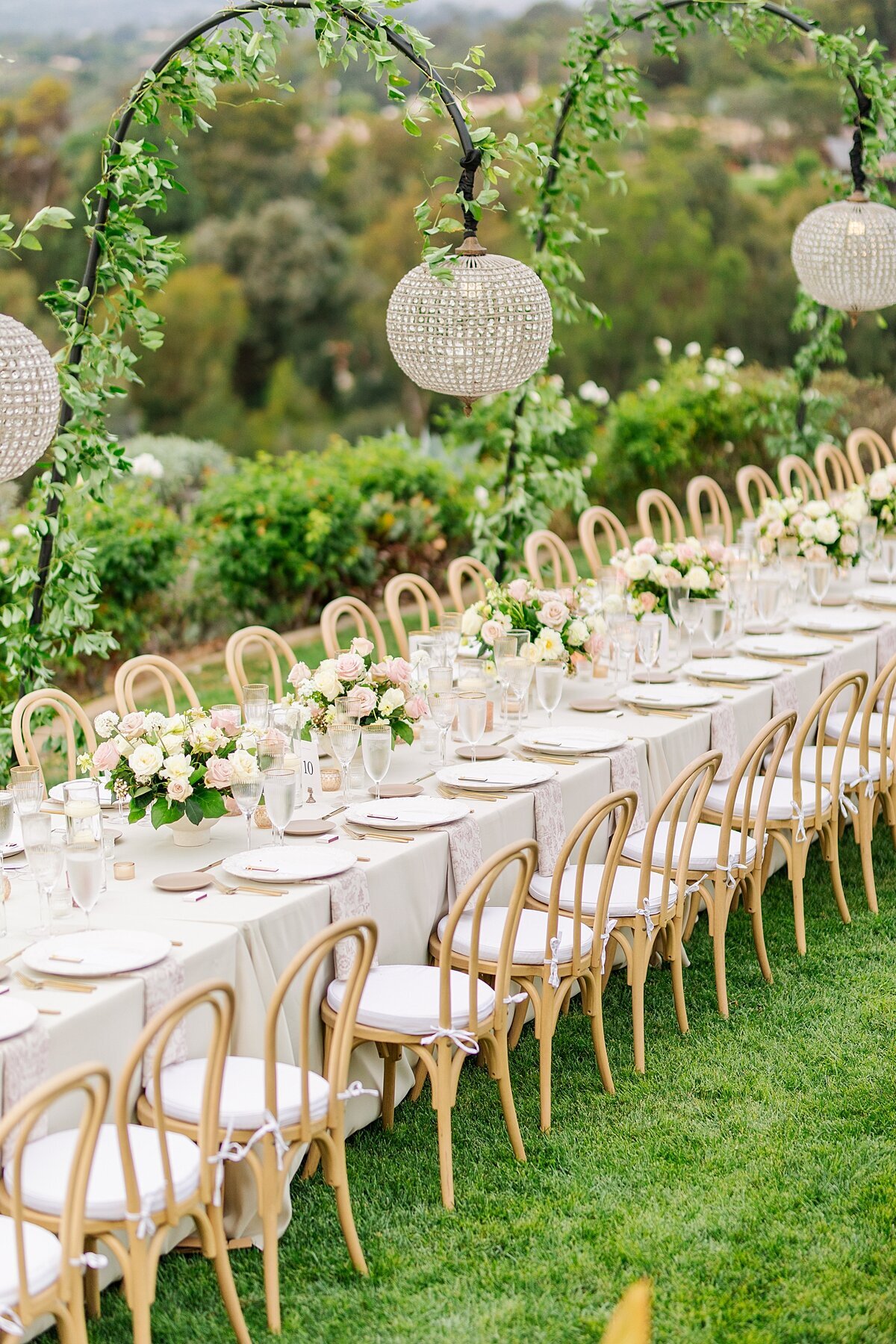 Long reception table with taupe linens and blush napkins and cutlery in front of ooden chairs with white seat covers with ivy-covered crystal chandeliers hanging overtop at Rancho Santa Fe private residence outdoor wedding reception.