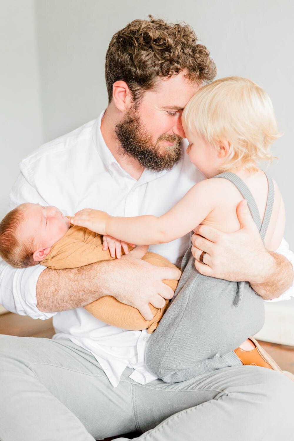 Dad snuggles toddler in one arm while the toddler gently pats the newborn in dad's other arm