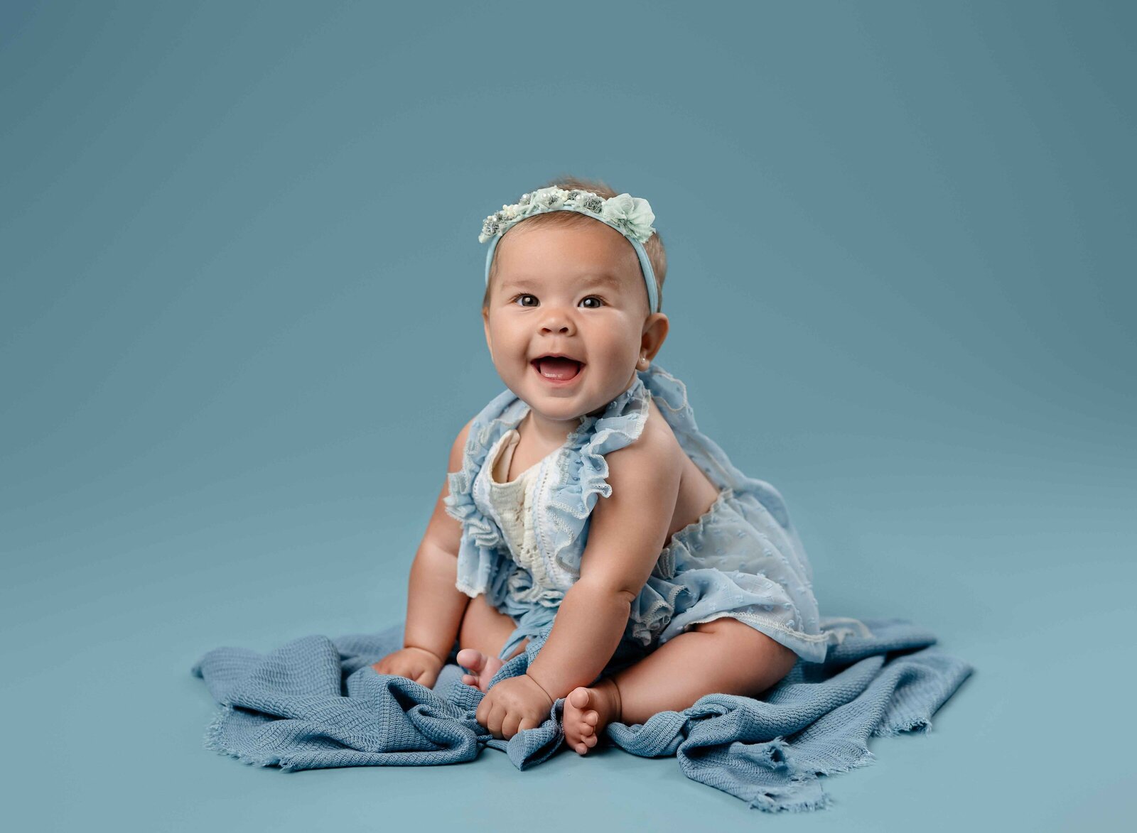 6 month baby girl sitting up with a blue outfit on and giving big smiles to the camera. Photo taken at Atlanta baby photo studio.