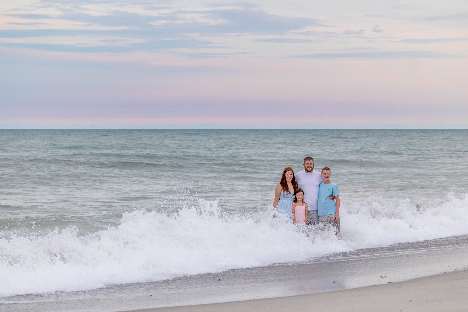 A family of four stands in the gentle waves at sunset, dressed in light pastel colors. The soft pink and blue sky reflects the serene atmosphere of a beach evening. Ideal for North Carolina family photography, capturing playful and scenic moments at the shore.