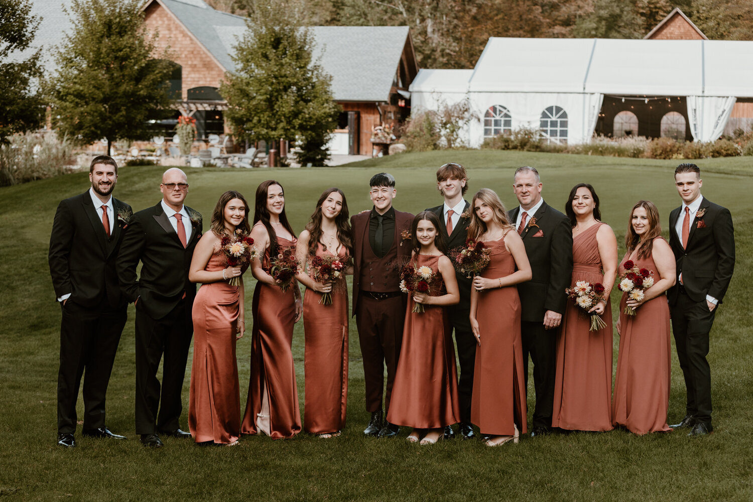 A group portrait of well-dressed individuals at a wedding. They are standing on a lush lawn with The Venue at Winding Hills, featuring a charming barn and white marquee tent, in the background.
