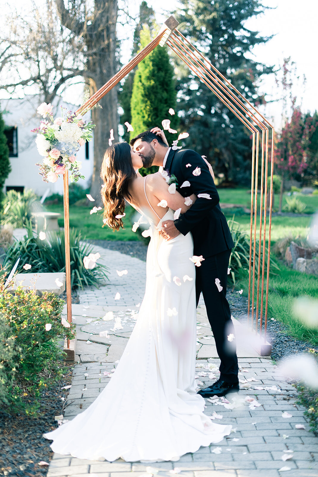 Ceremony kiss with flower petals and brass arch at Cottage at Riverbend Wedding by the Best Boise Wedding Photographers