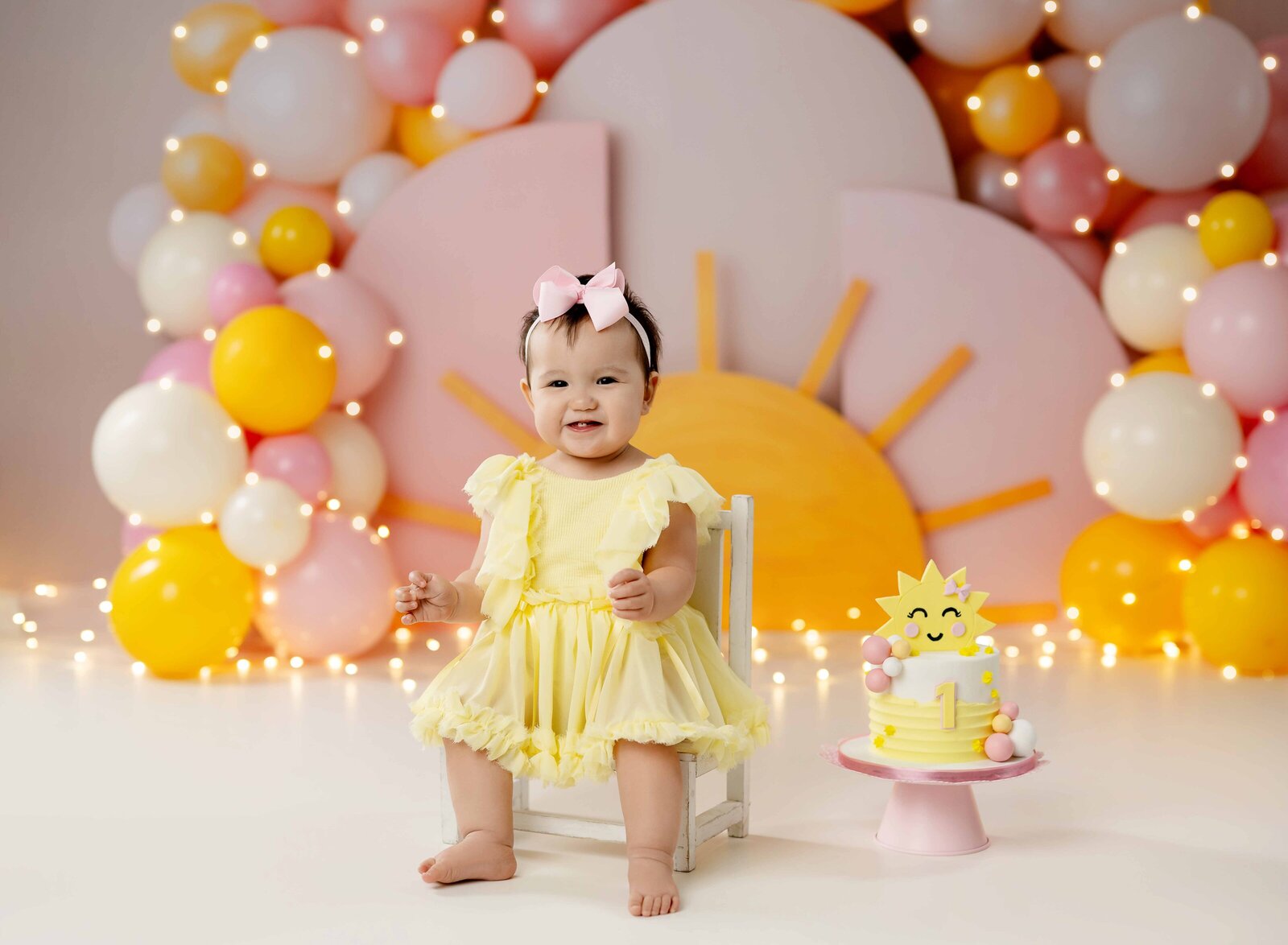 you are my sunshine birthday set with pink and yellow balloons and sunshine rays. Baby in yellow dress sitting on stool and smiling.