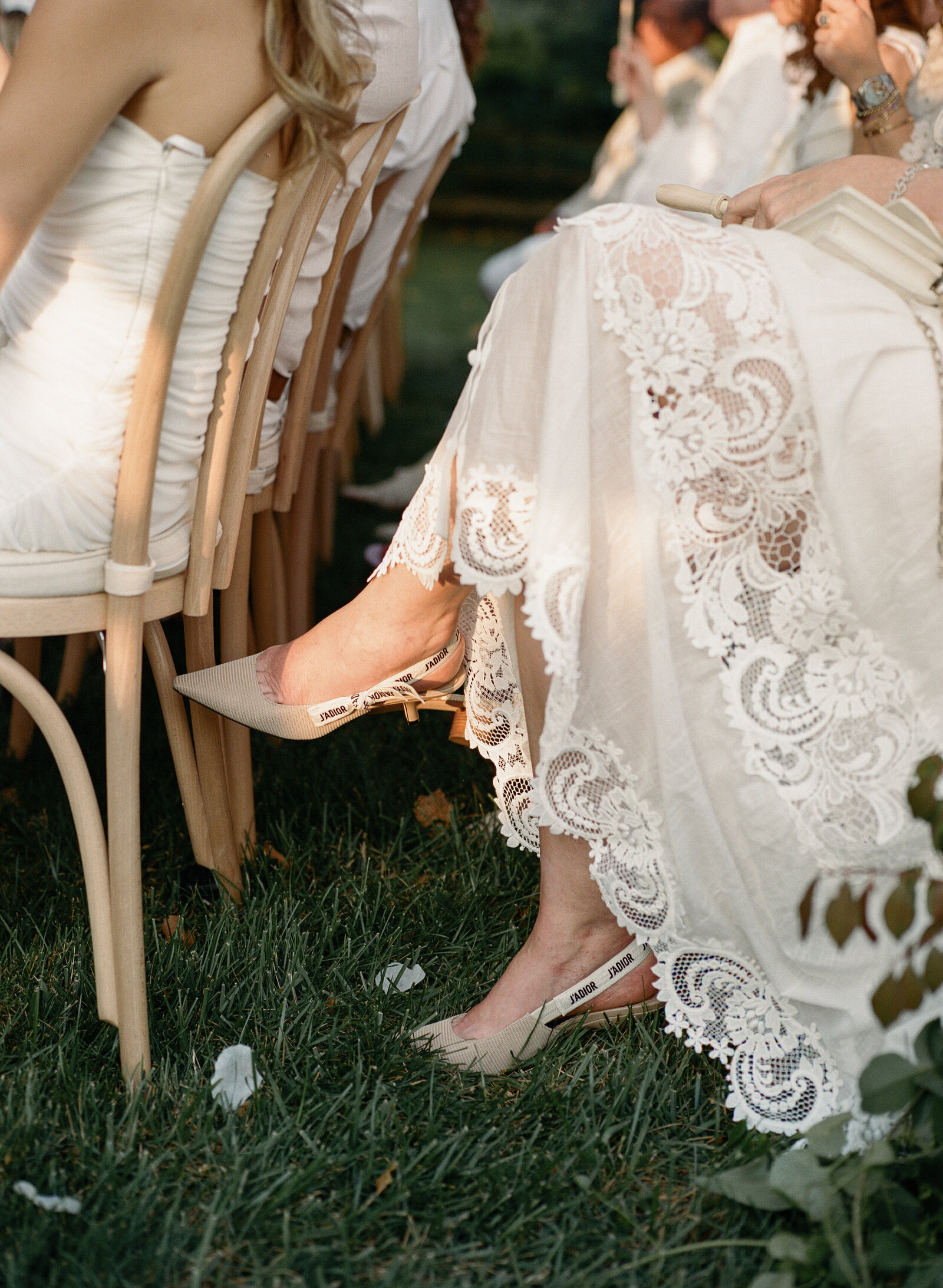 Close up detail of wedding guest's feet sitting cross legged at outdoor ceremony wearing white lace trimmed dress and tan Dior pumps