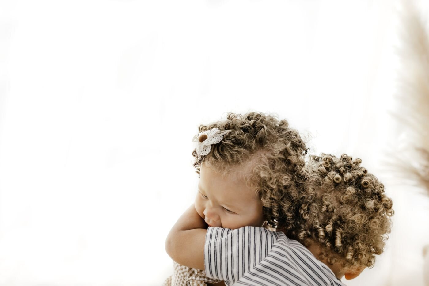 A tender close-up of two young children with beautiful curly hair embracing each other against a bright, soft background. A sweet sibling moment