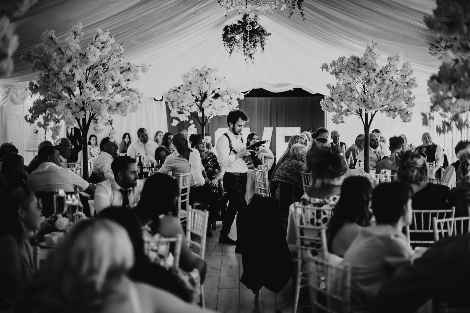Tom Keenan photographing a wedding speech at a reception, surrounded by guests in a beautifully decorated venue.