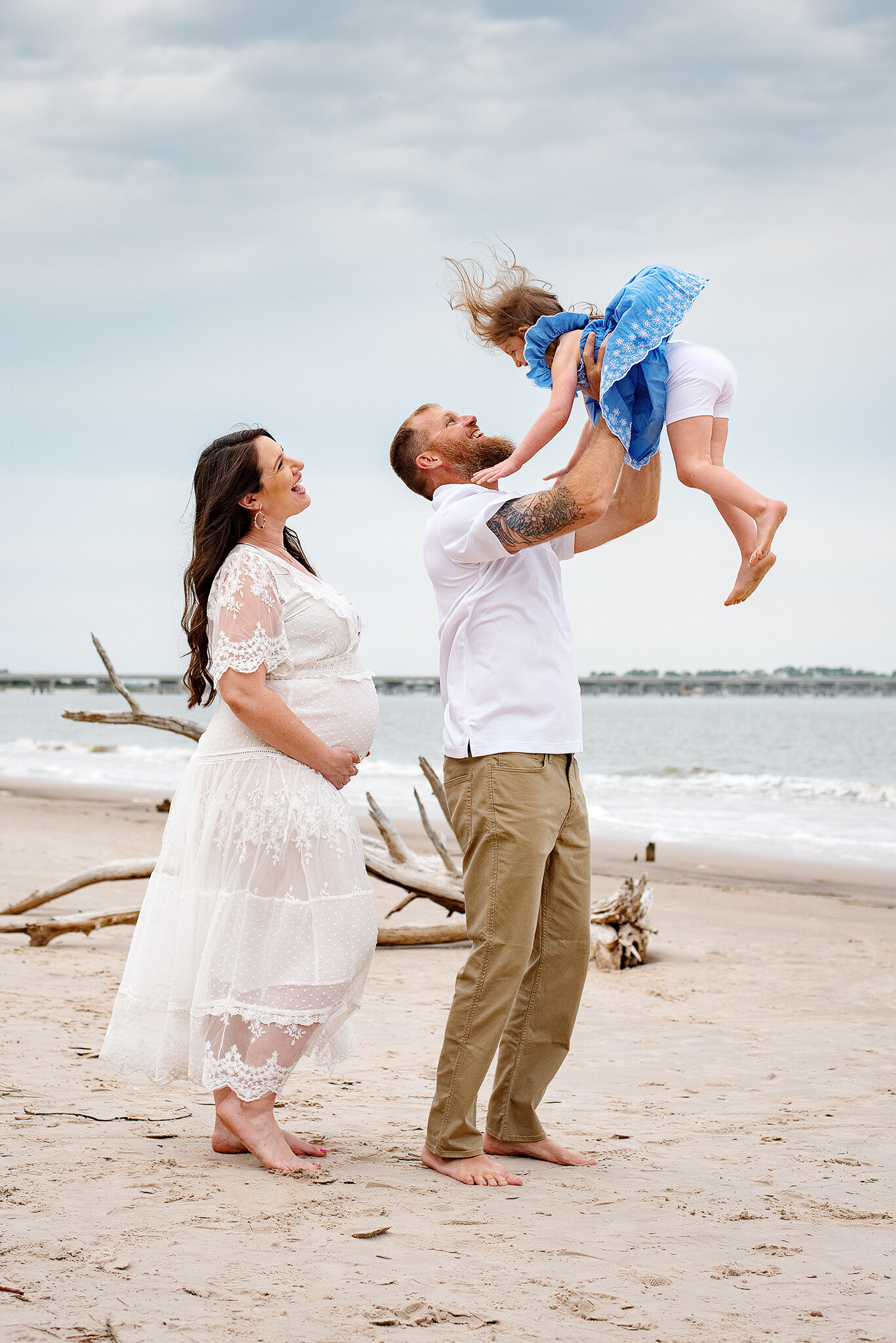 Dad lifts daughter in blue dress while pregnant mom in white watches at Talbot Island in FL.