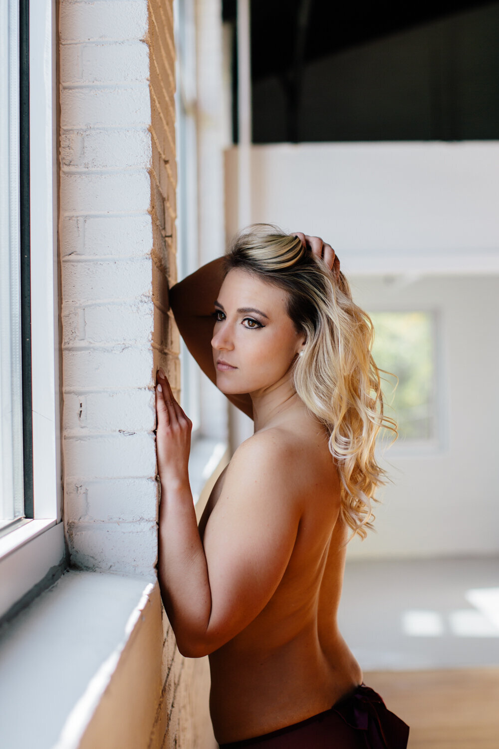 Blonde hair girl facing a white brick wall looking at a window without a shirt captured by boudoir by Steph