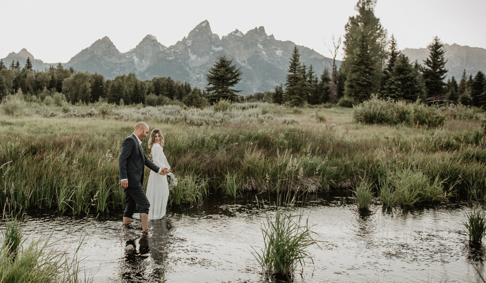 jackson hole wedding photographers captures bride and groom walking through the river at schwabacher landing as they hold hands with a clear view of the mountains for their grand teton wedding