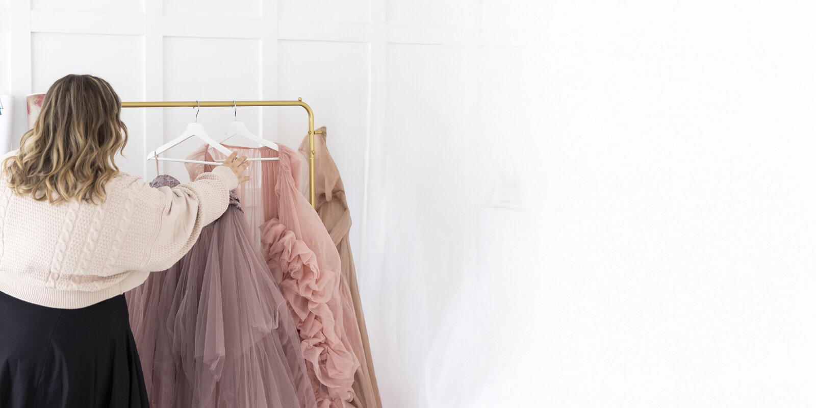 A woman with wavy hair and a beige sweater selects a pink tulle dress from a gold clothing rack, conveying a sense of elegance and choice.