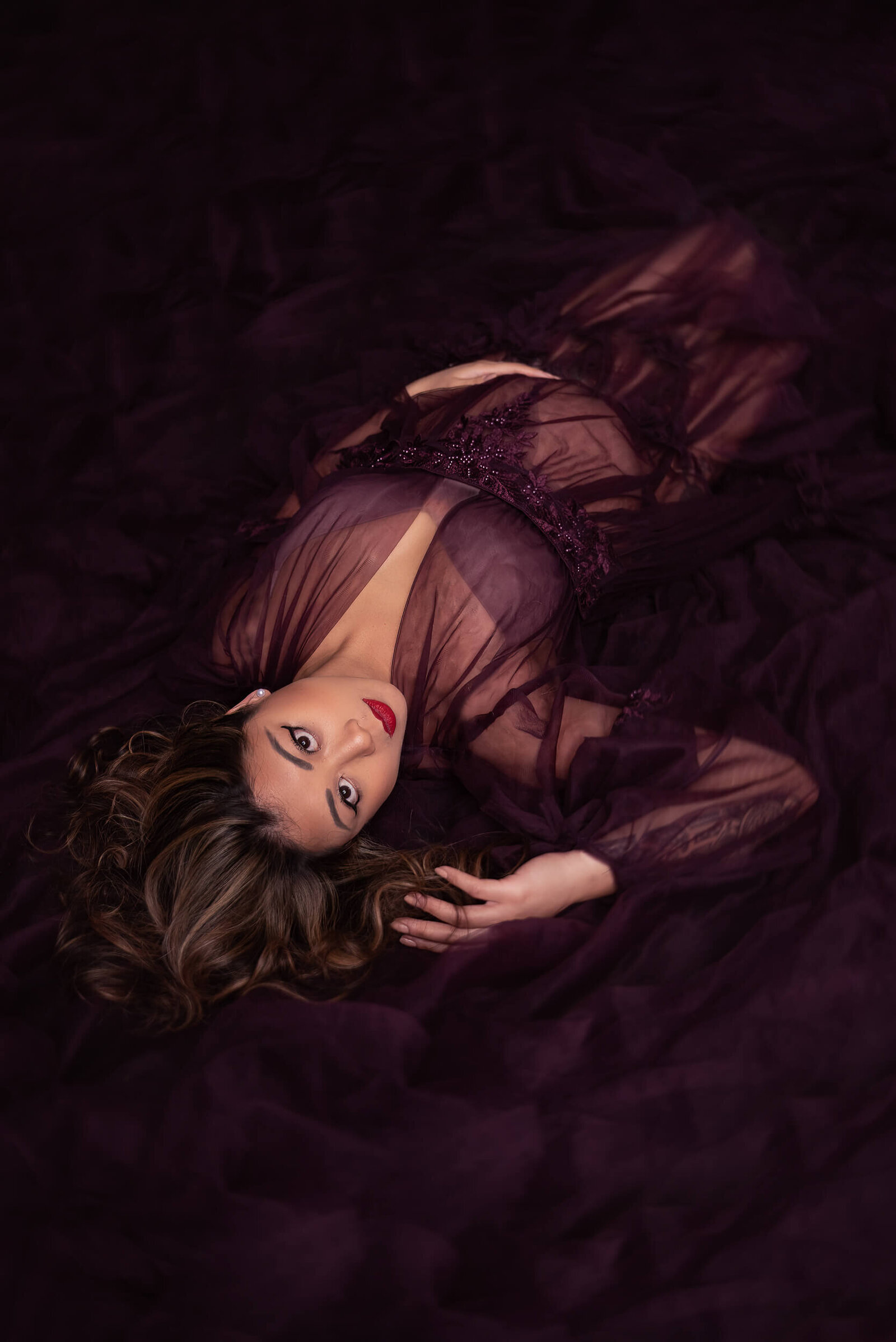 An expectant mother lies on a rich plum fabric backdrop, gazing softly at the camera, exuding confidence and serenity.