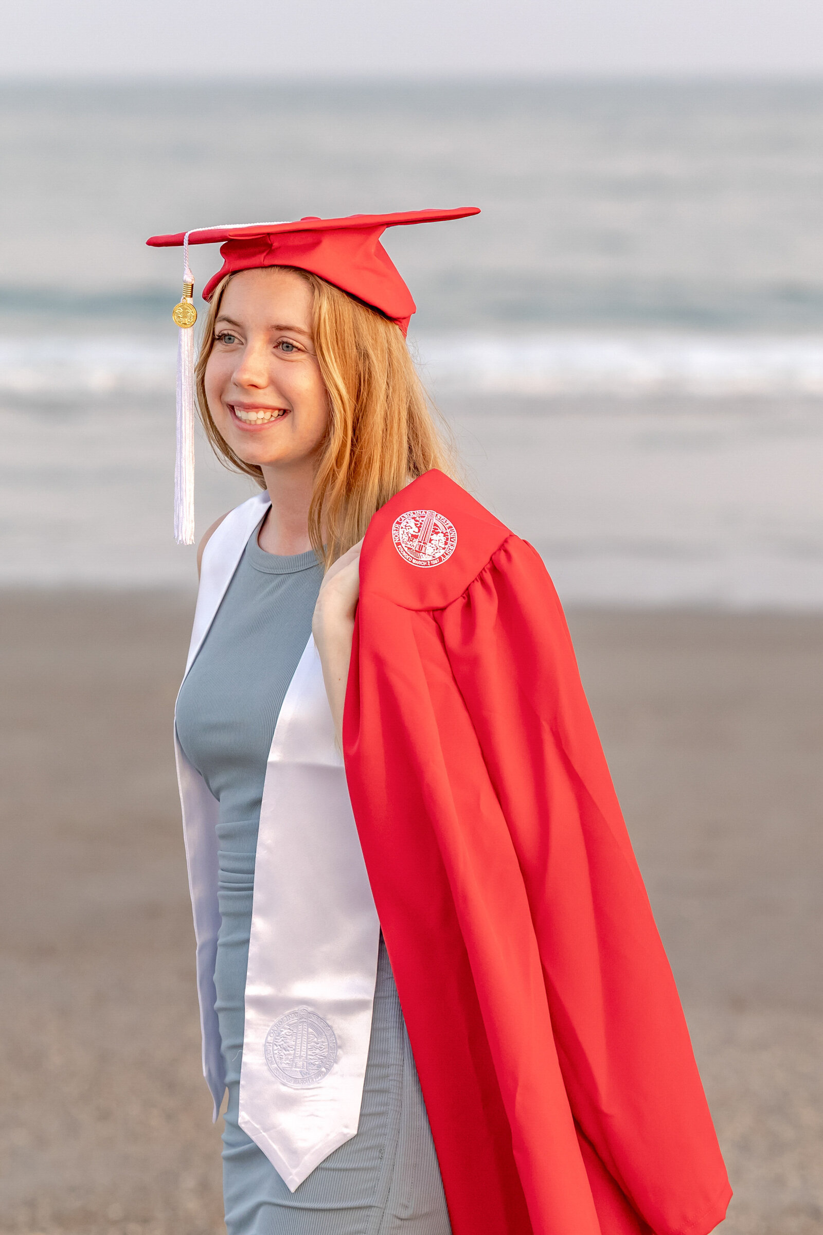A college senior in a red cap and gown smiling by the ocean in Ocean Isle Beach, North Carolina. This vibrant portrait highlights the joy of graduation, perfect for capturing memorable senior moments on the beach.