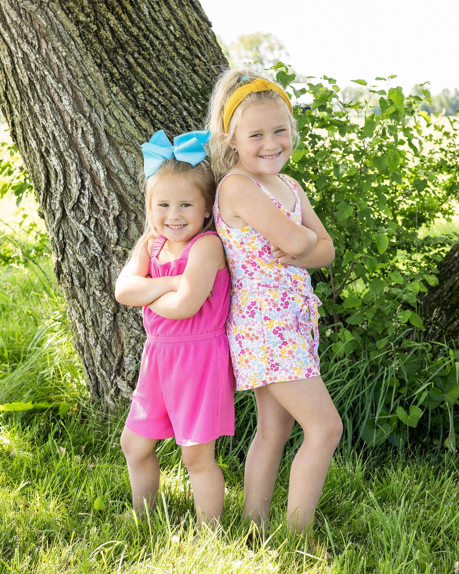 Two young girls standing back to back in front of a tree.  One has blonde hair wearing a blue bow and a pink romper with arms crossed, smiling at the camera.  The other girl has blonde hair wearing a white flower pattern romper with arms crossed, smiling at the camera.