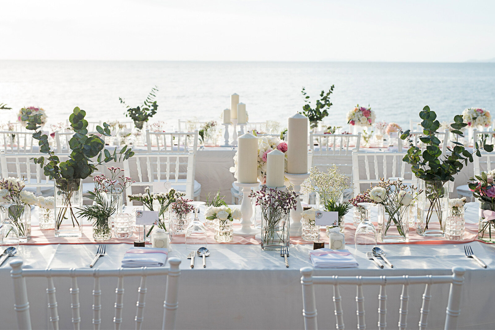 Wedding reception table with dinnerwares, floral and candle centerpieces with white chairs, and an ocean view