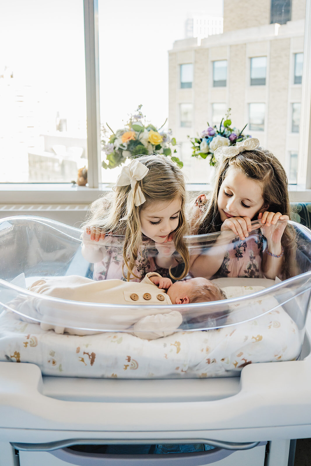 big sisters peek over side of hospital bassinet at new baby brother