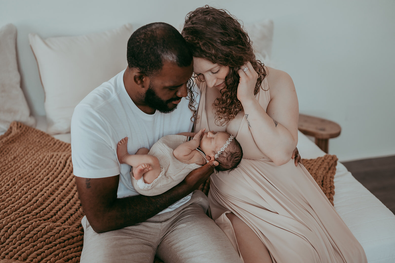 husband and wife sit on bed in neutral wardrobe holding their newborn