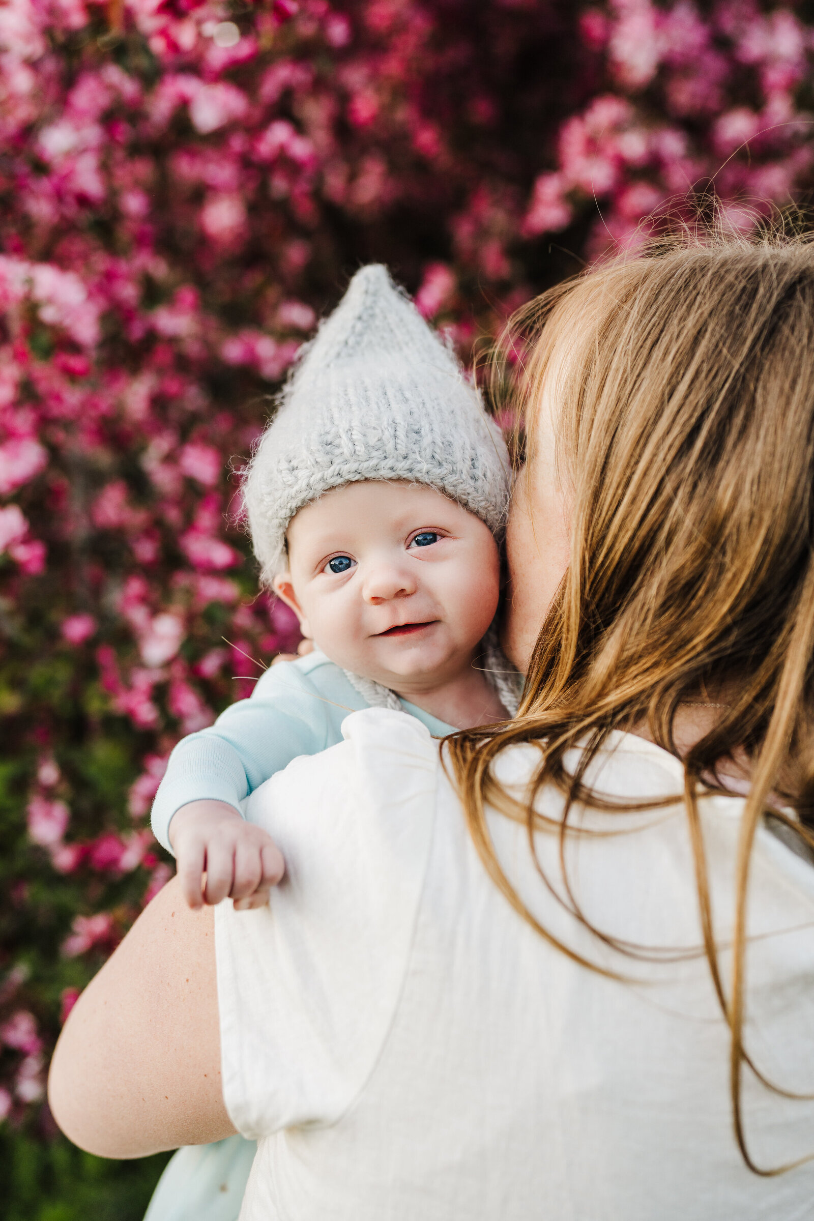 baby in wool hat looks over moms shoulder with blossoming trees