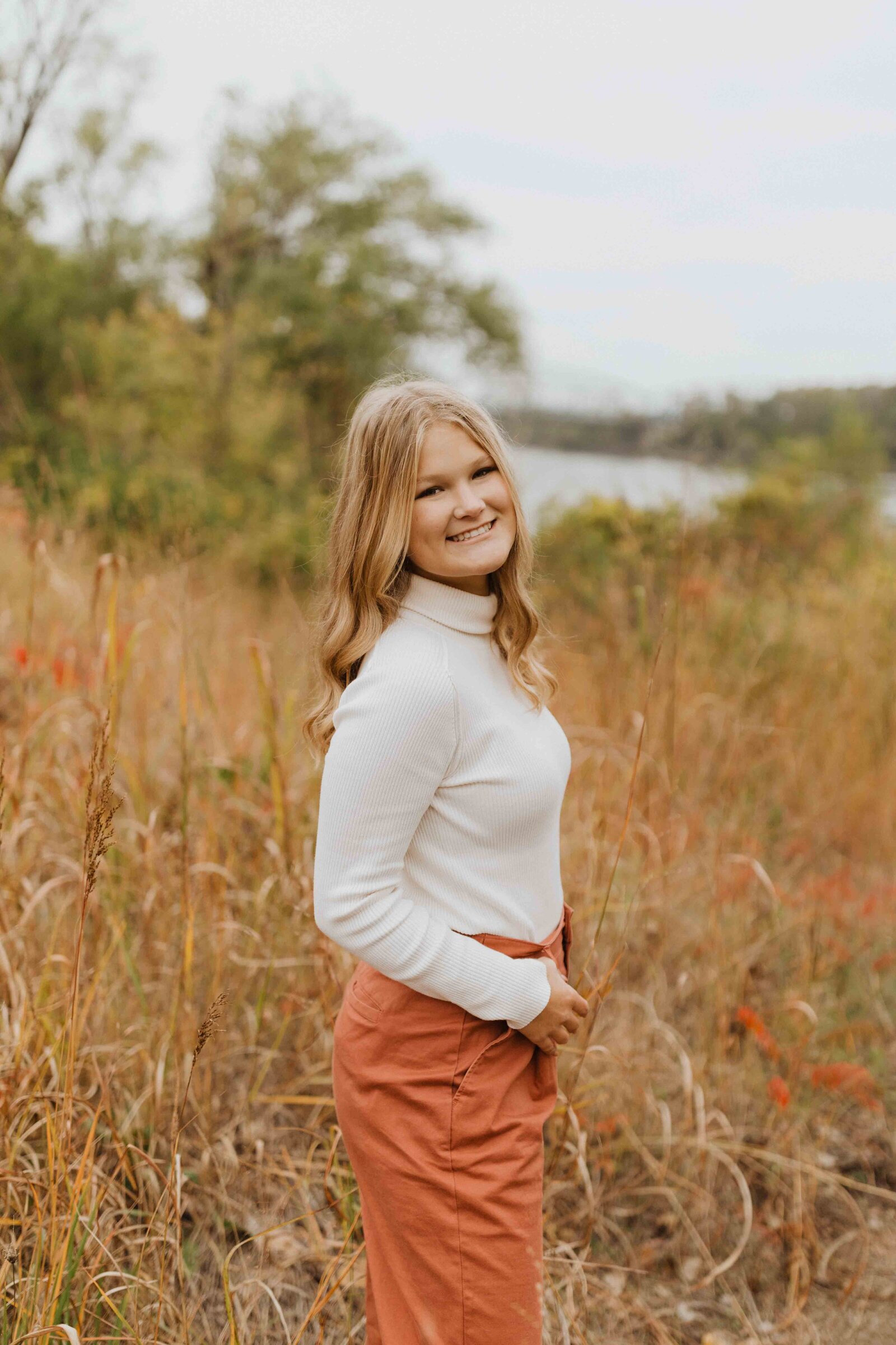 Girl is standing in the middle of golden field. She has her thumb through belt loop, looking at camera and smiling.